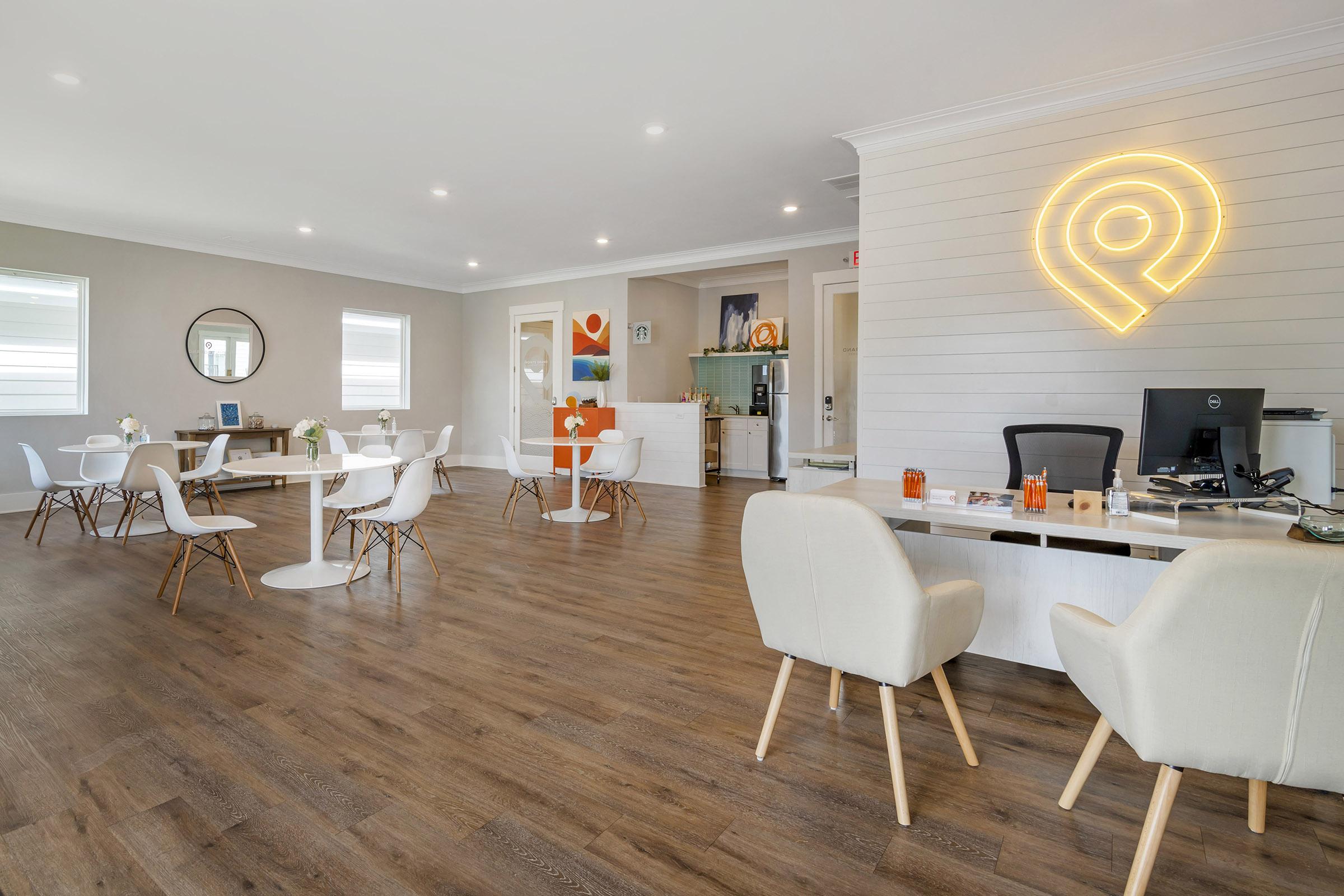 Interior of a modern office space featuring light-colored walls and wooden flooring. There are several white tables with chairs arranged neatly, a reception desk with a computer, and decorative elements on the walls. Soft lighting enhances a welcoming atmosphere.