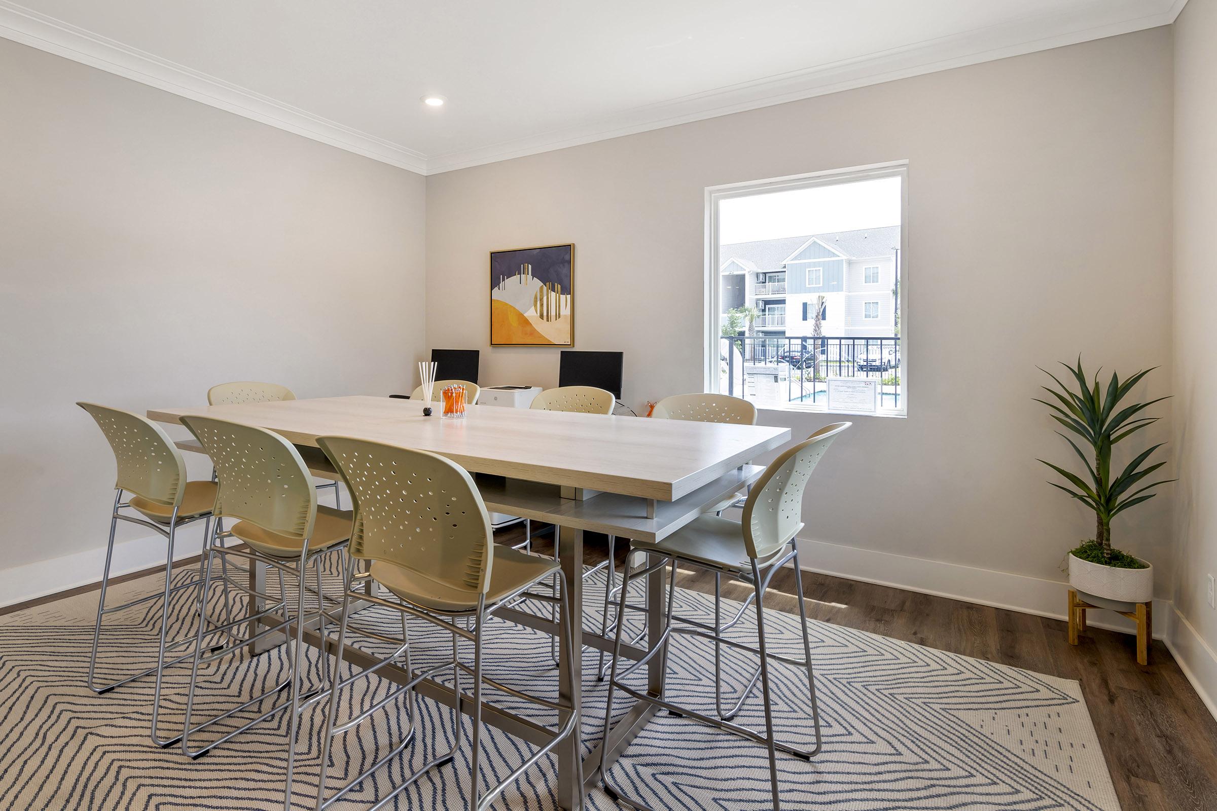 A modern dining area featuring a large wooden table surrounded by beige chairs. A window lets in natural light, overlooking an outdoor view. The room has neutral-toned walls and a stylish rug beneath the table, complemented by a potted plant in the corner.