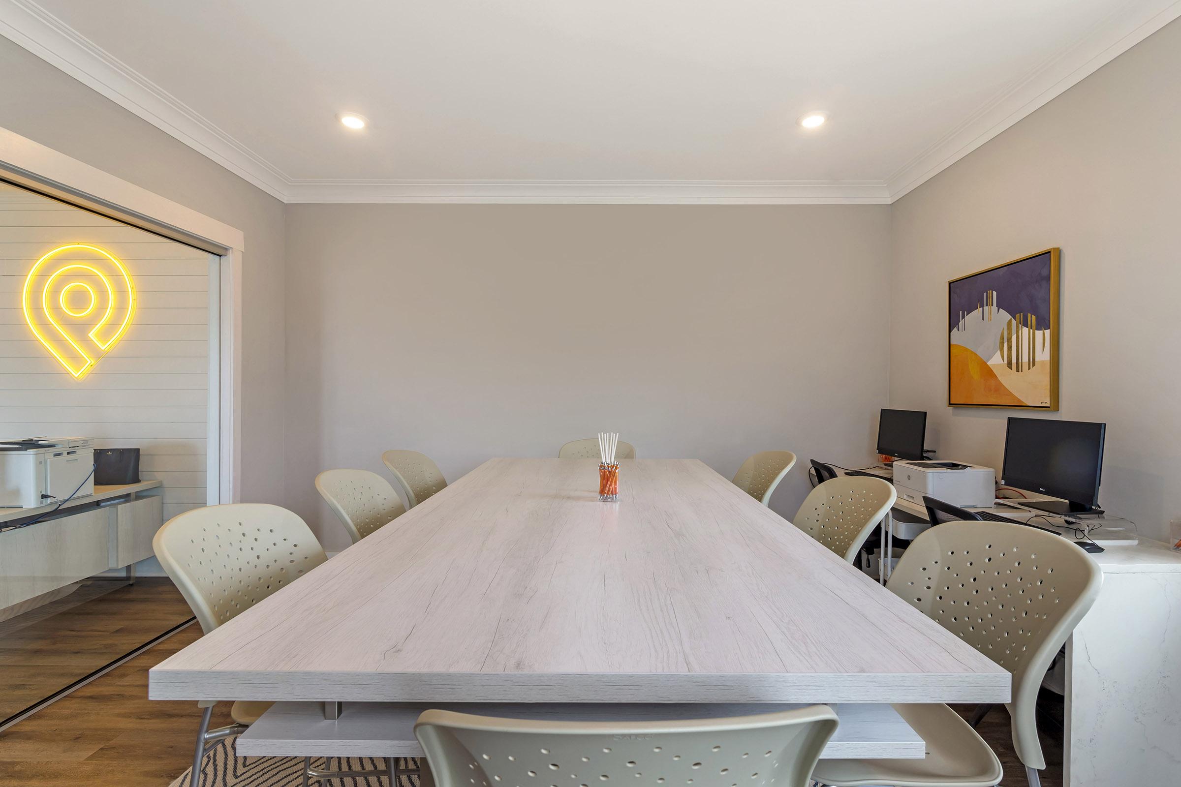 A spacious meeting room featuring a large wooden table surrounded by light-colored ergonomic chairs. There are two computers on desks against the wall. A piece of abstract art hangs on the wall, and a decorative element resembling a location pin is visible in the background. The room has neutral-colored walls and natural light.