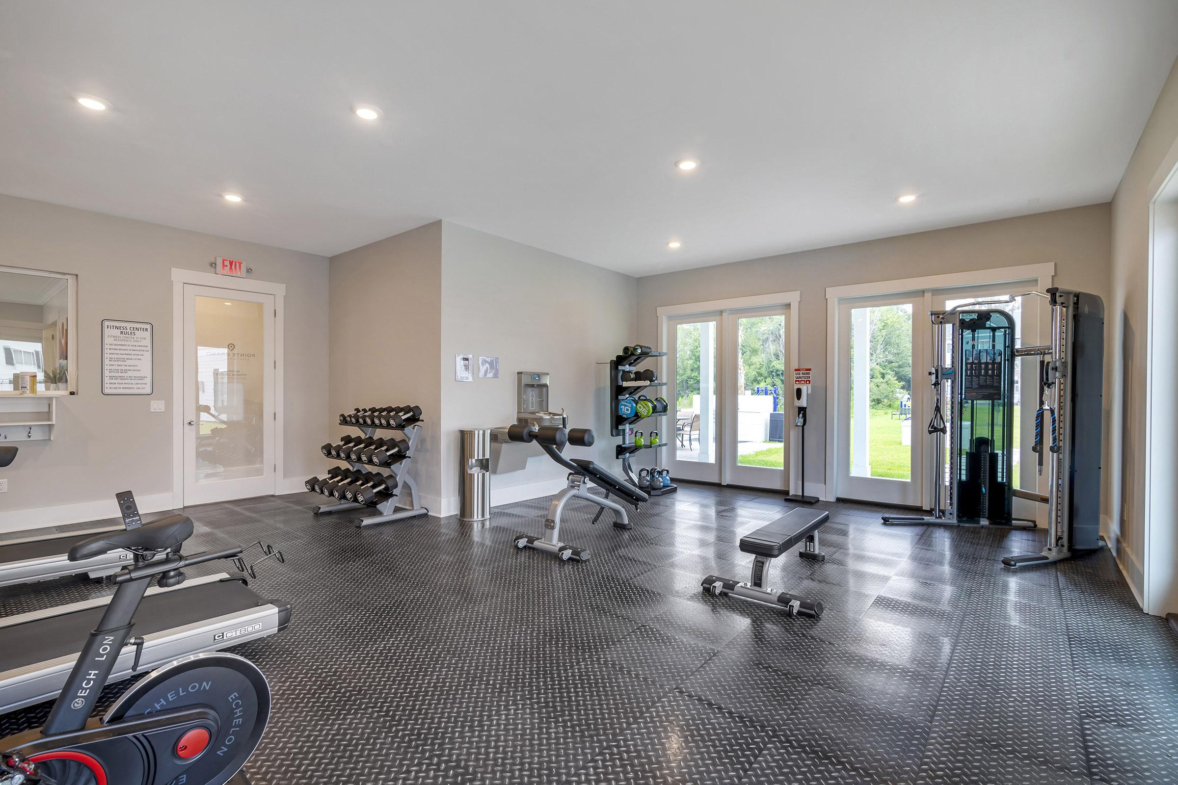A modern, well-lit gym featuring various fitness equipment. The space includes dumbbells on a rack, a bench press, and cardio machines like a stationary bike. Large windows provide natural light and a view of the outdoors, creating an inviting atmosphere for workouts.