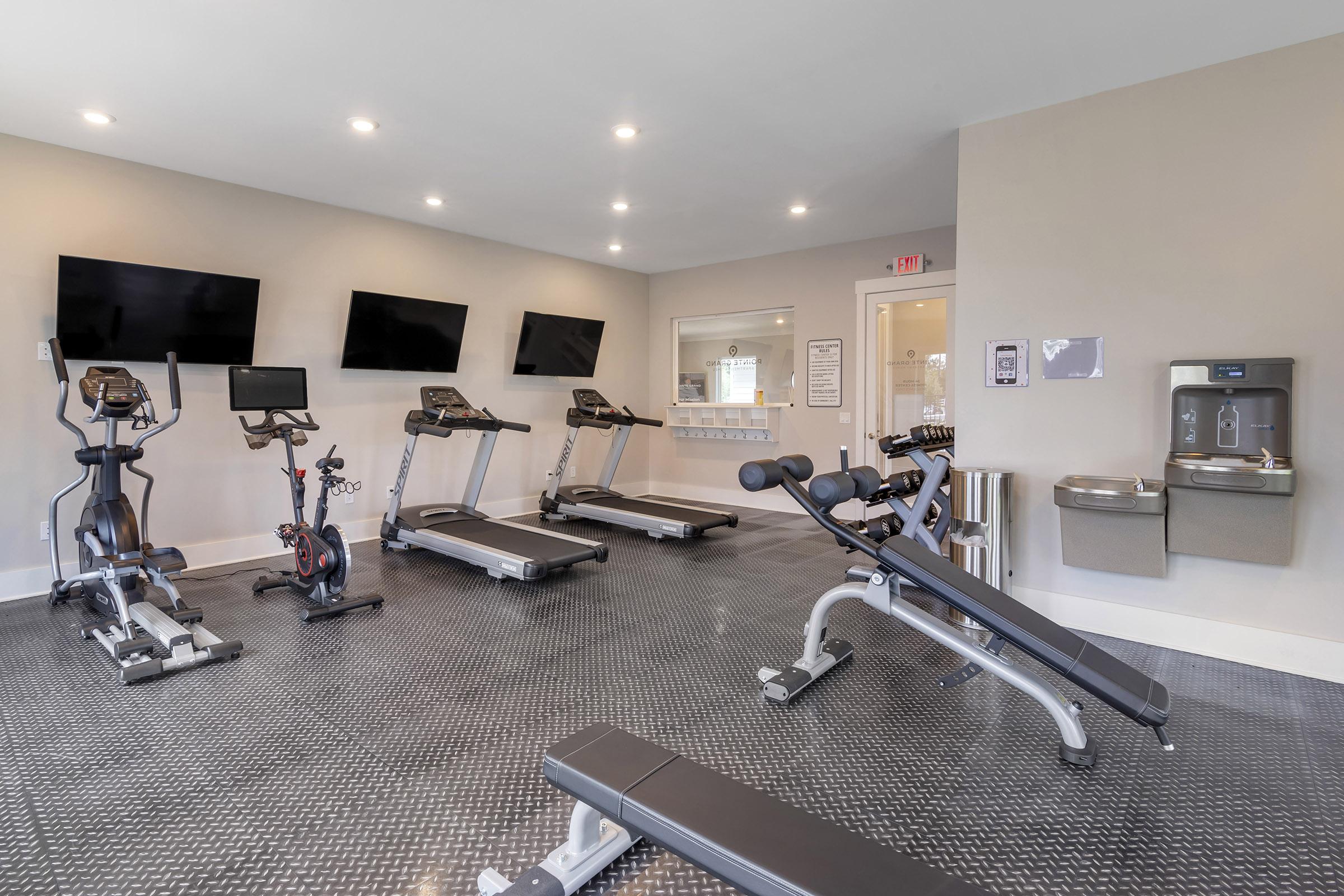 A modern gym interior featuring multiple exercise machines, including treadmills, a stationary bike, and a weight bench. There are three wall-mounted televisions, a large mirror, and a water fountain. The flooring is textured and dark, contributing to a sleek workout environment.