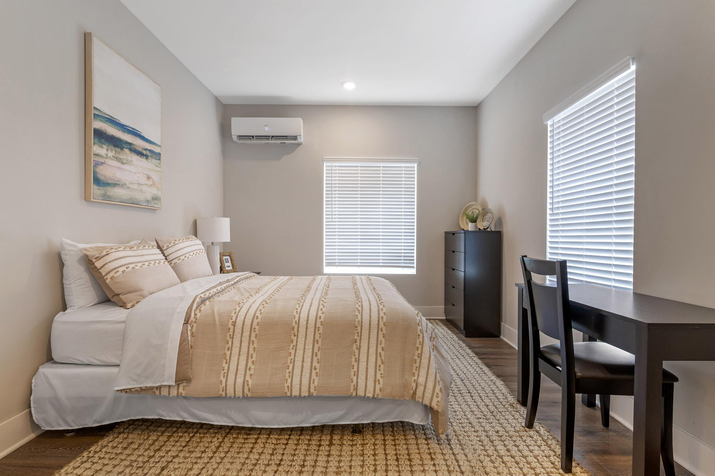 A cozy bedroom featuring a neatly made bed with a patterned beige and white comforter. There is a small nightstand with a lamp beside the bed. A modern black dresser stands against one wall, and a simple black desk and chair are positioned near a window with white blinds. A piece of abstract art hangs on the wall.