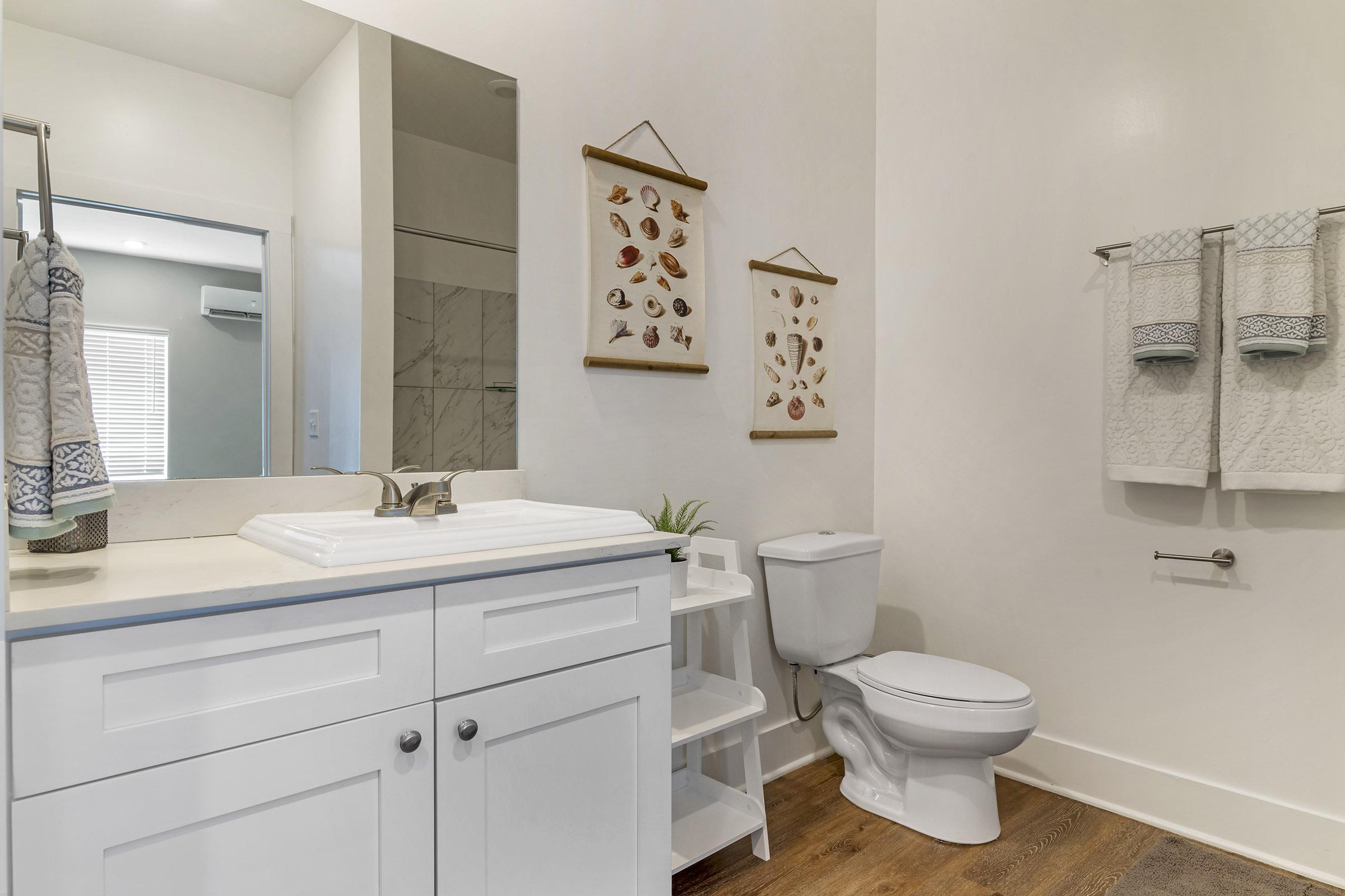 A clean, modern bathroom featuring a white vanity with a sink, decorative wall art displaying botanical prints, plush towels on a rack, and a toilet. Natural light filters in through a window, enhancing the bright, minimalist design.
