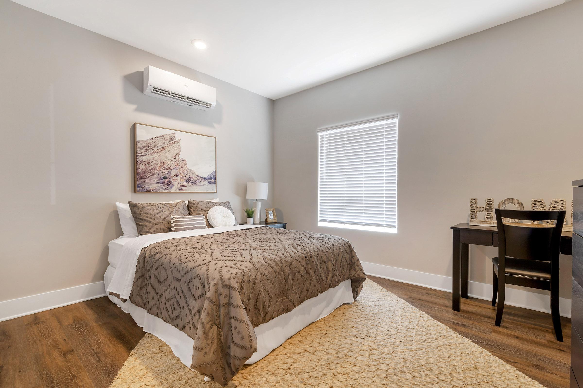 A cozy bedroom featuring a neatly made bed with a patterned brown blanket, a nightstand with a lamp, and a small desk with a black chair. Natural light filters through a window with white blinds, and a textured area rug enhances the warm atmosphere of the space.