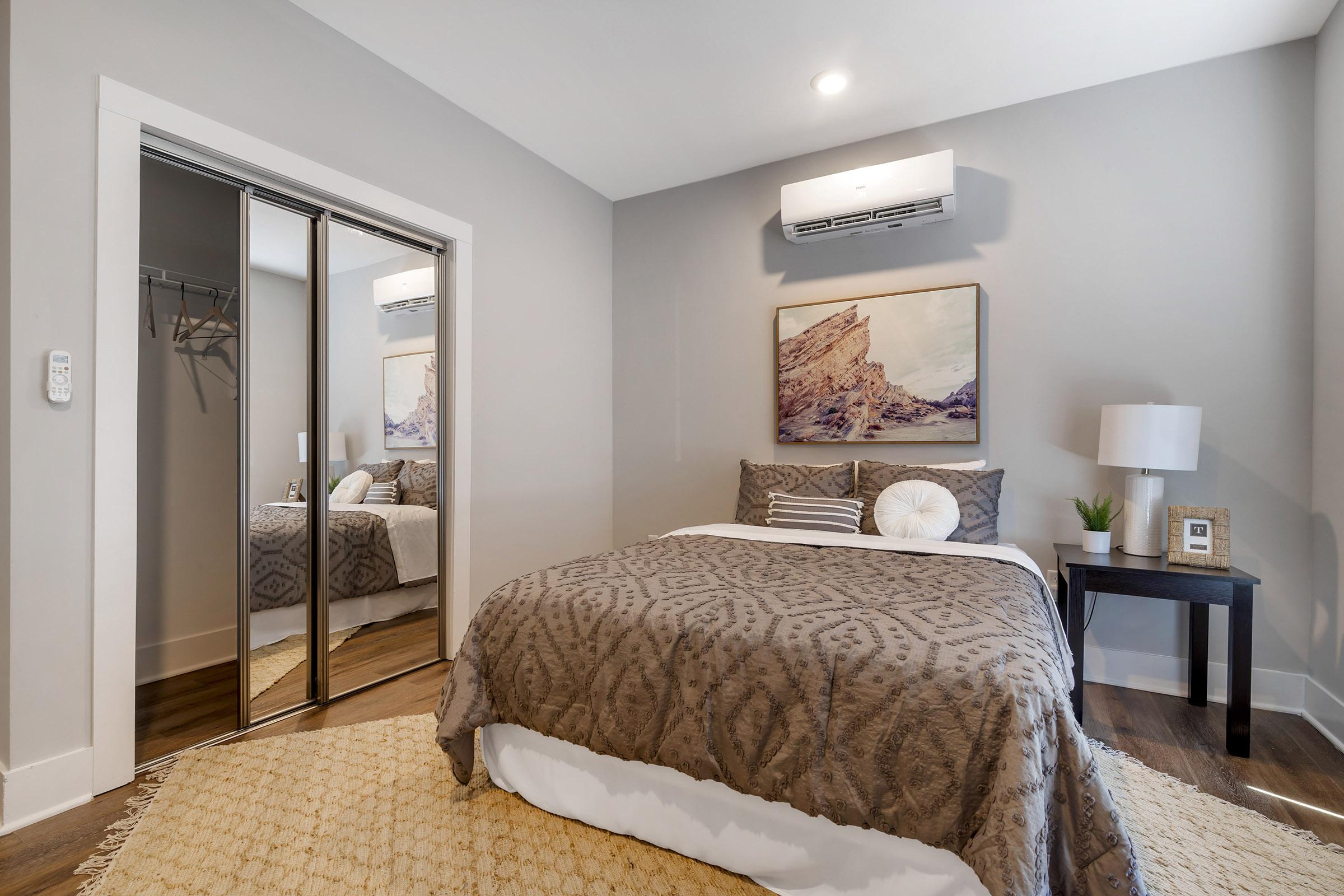 A cozy bedroom featuring a queen-sized bed with a patterned brown comforter, a bedside table with a lamp, and a decorative picture on the wall. A large mirror closet reflects the room, and there's a small plant beside the bed. The walls are painted a soft gray, providing a calming atmosphere.