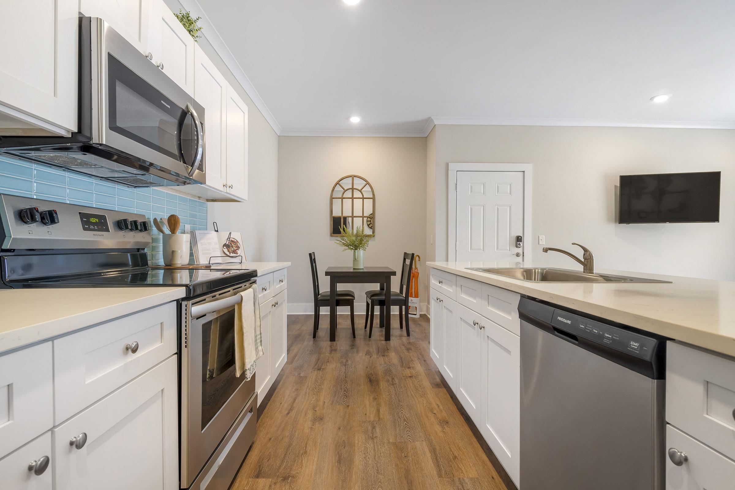 Modern kitchen with white cabinetry, stainless steel appliances including an oven, microwave, and dishwasher. A small dining area with a table and chairs is visible. Light blue tile backsplash and wooden flooring enhance the bright, inviting atmosphere. Natural light fills the space from a nearby window.