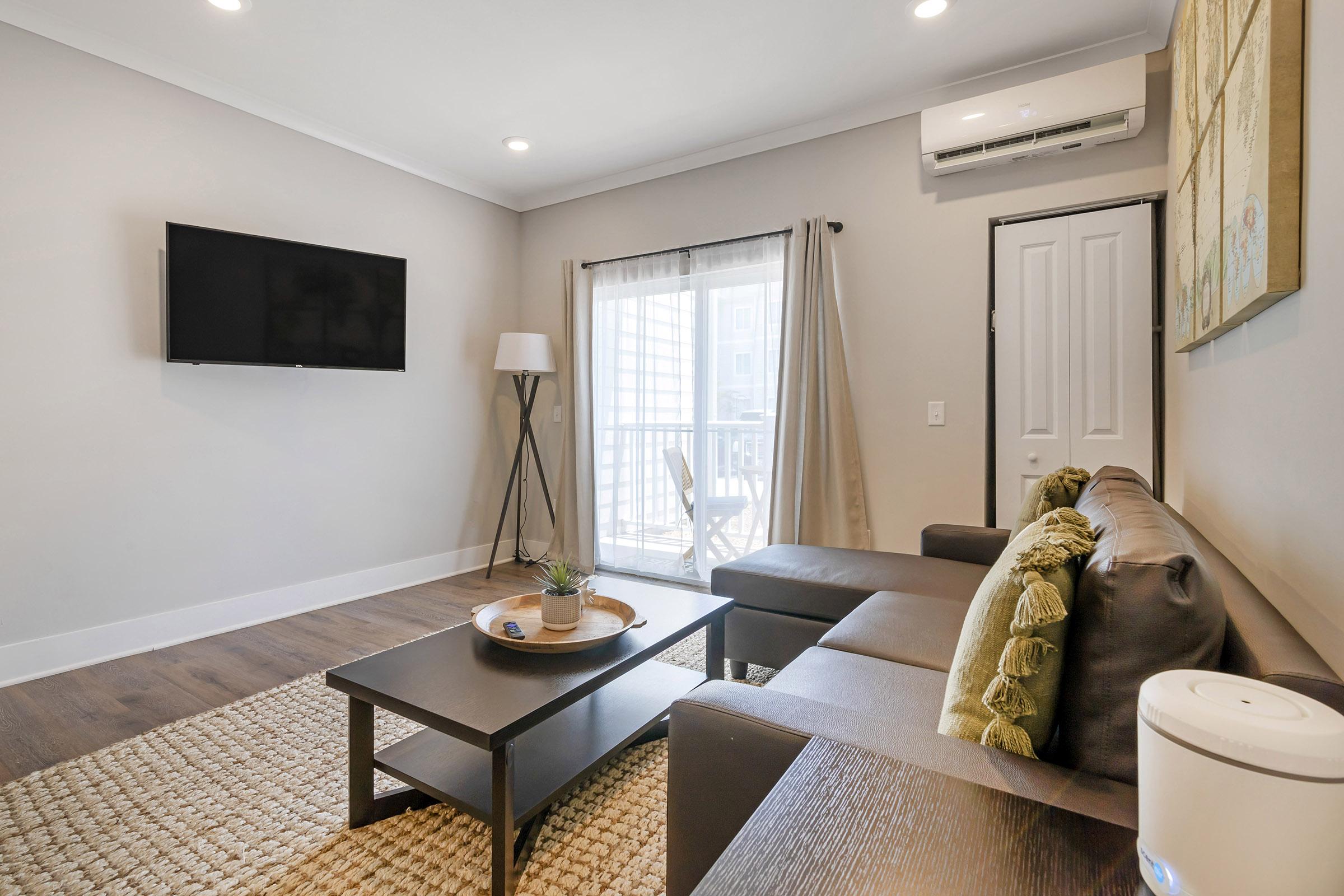 A cozy living room featuring a modern sofa with decorative cushions, a wooden coffee table with a small plant, and a rug. There’s a flat-screen TV mounted on the wall, a floor lamp, and large windows with light curtains, allowing natural light to fill the space. Air conditioning unit visible above the door.