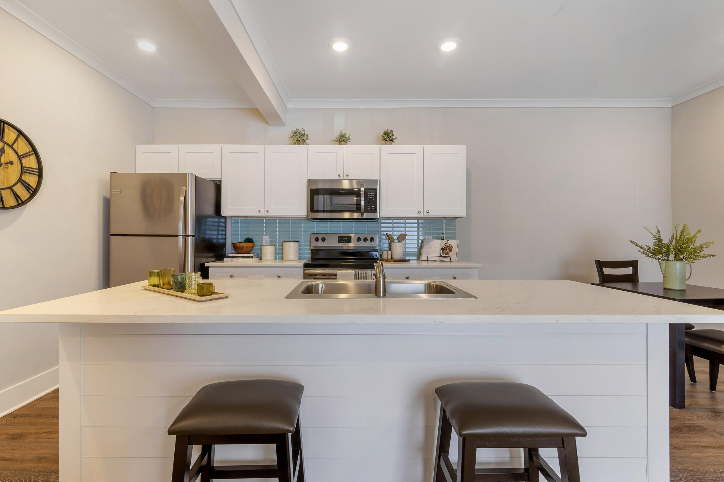 Modern kitchen featuring a large white island with two dark wood stools, stainless steel appliances, and a tiled backsplash. The space includes white cabinetry, decorative plants, and a dining area with a wooden table and chairs, illuminated by overhead lights.