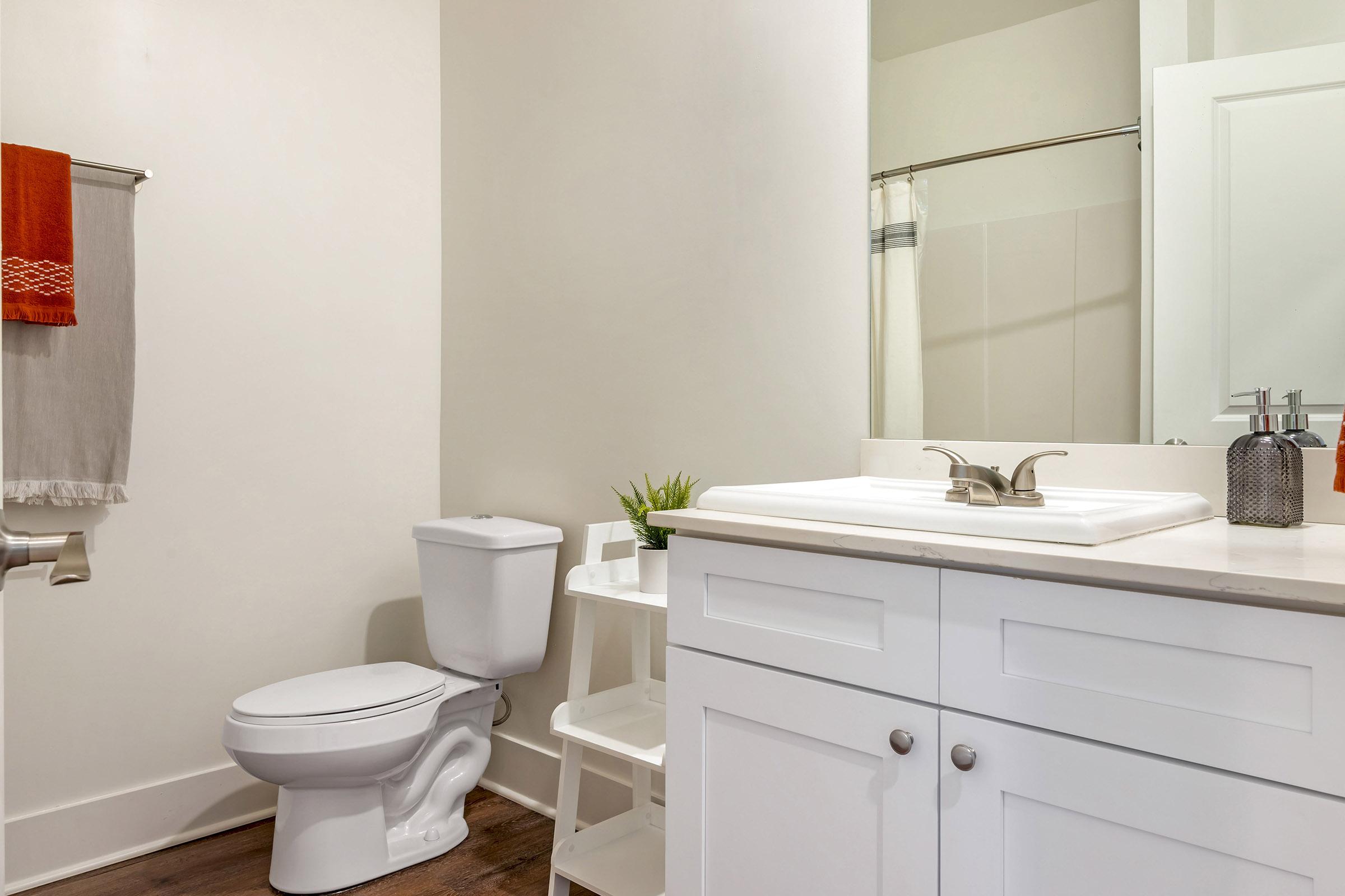 A clean and modern bathroom featuring a white toilet, a sink with a countertop, and cabinets below. There is a shower in the corner, and decorative items like a small plant and an orange towel add a pop of color to the neutral decor. Natural light illuminates the space, creating a bright atmosphere.