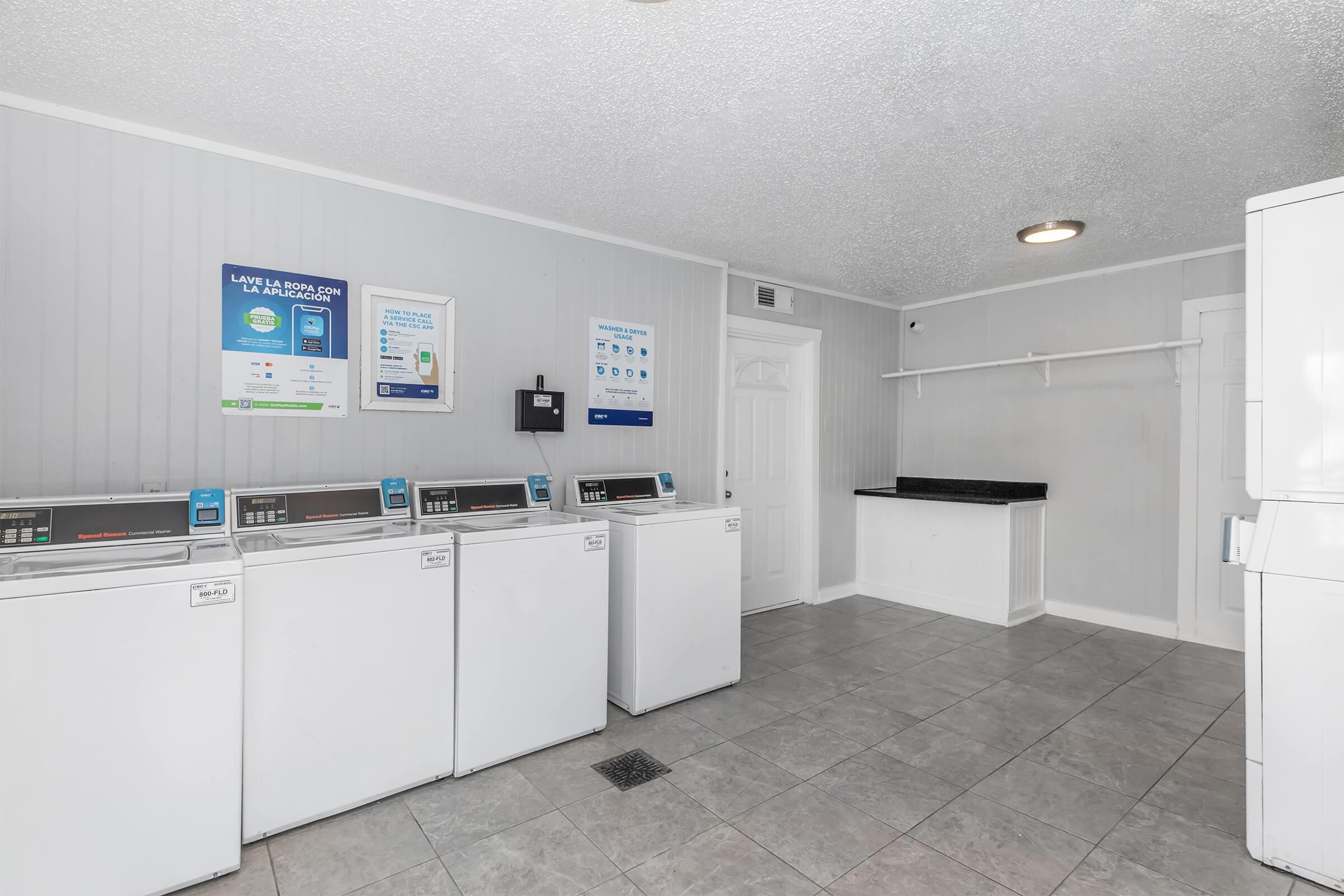 A clean, modern laundry room featuring several white washing machines along one wall. The space has light gray walls, tiled flooring, a counter area, and informational posters about laundry services. Bright lighting enhances the overall cleanliness and functionality of the room.