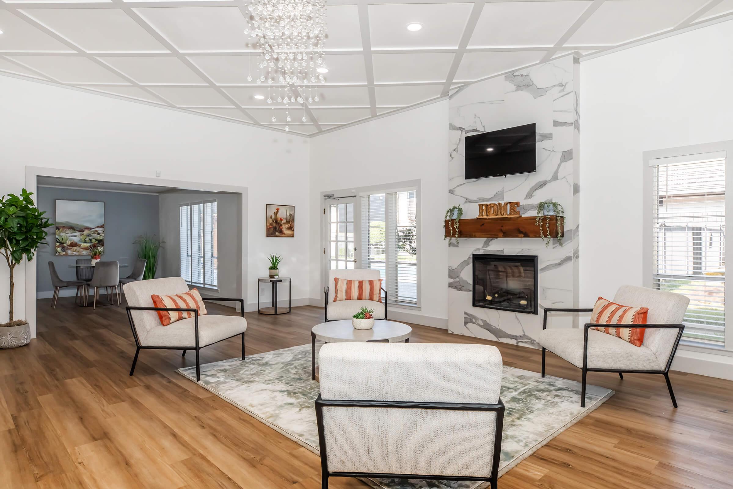 Cozy living room featuring four modern chairs around a circular coffee table, with a decorative plant, a stylish rug, and a wall-mounted television above a marble-accented fireplace. Bright, airy space with large windows, wood flooring, and elegant lighting fixtures.