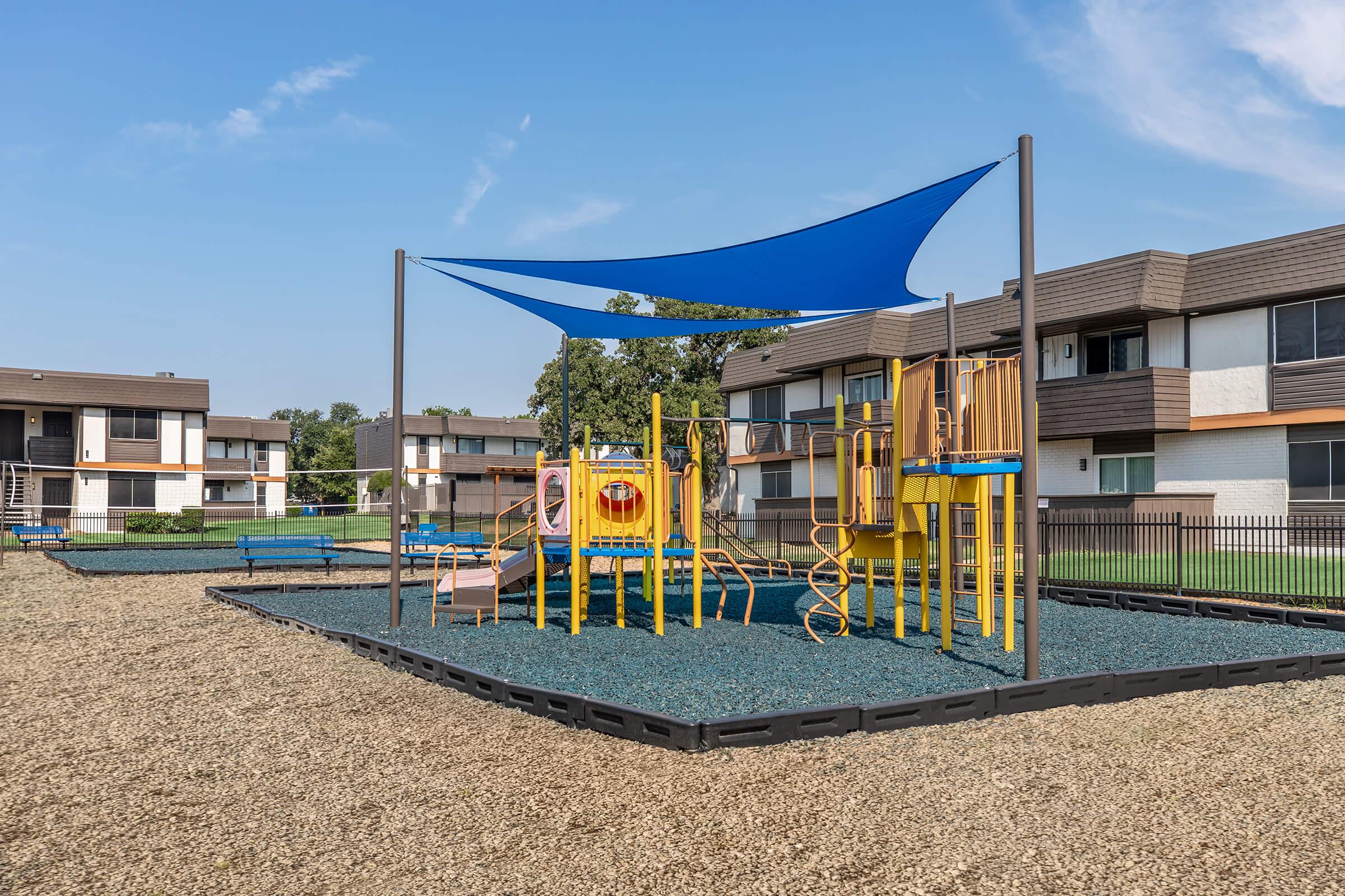 Children's playground featuring a yellow play structure with slides and climbing features, shaded by a blue canopy, situated in a gravel area. In the background, there are residential buildings and a grassy space, indicating a community setting. Clear blue sky above.