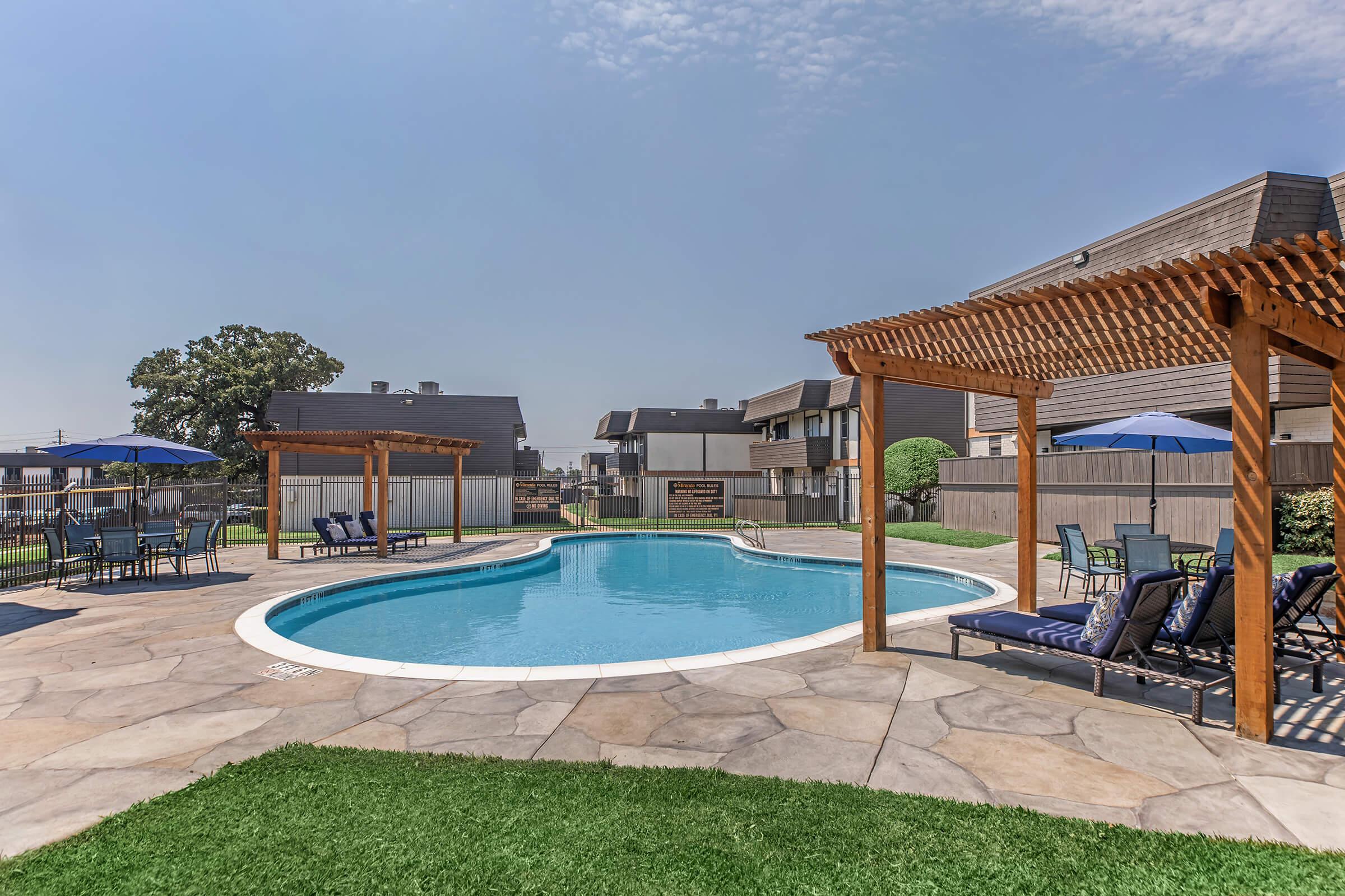 A swimming pool surrounded by a paved deck, featuring two wooden pergolas with outdoor seating. There are several lounge chairs and umbrellas near the pool, with grassy areas and apartment buildings in the background under a clear blue sky.