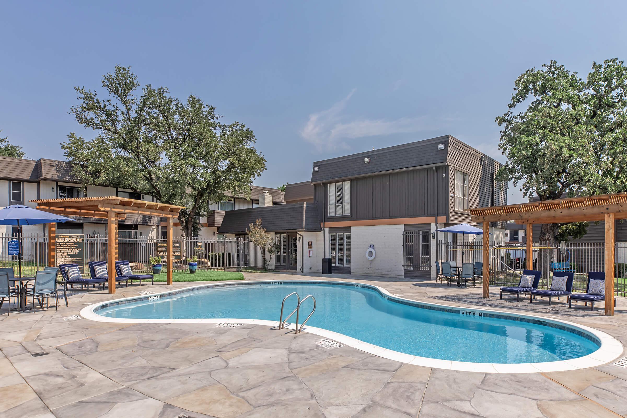 Outdoor swimming pool with a curved shape surrounded by lounge chairs and tables. There are two wooden pergolas providing shade, and the area features well-maintained grass and trees. In the background, a modern two-story building is visible, set against a clear blue sky.