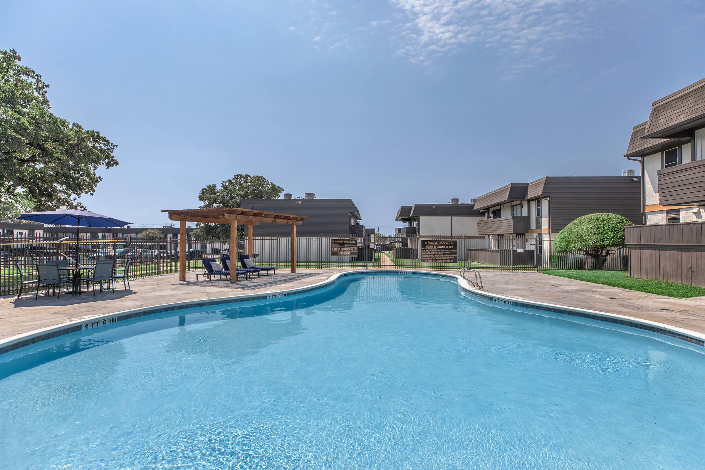 A clear swimming pool surrounded by a patio area with lounge chairs and tables. In the background, there are two modern apartment buildings and trees. The sky is bright with some clouds, creating a sunny outdoor atmosphere.