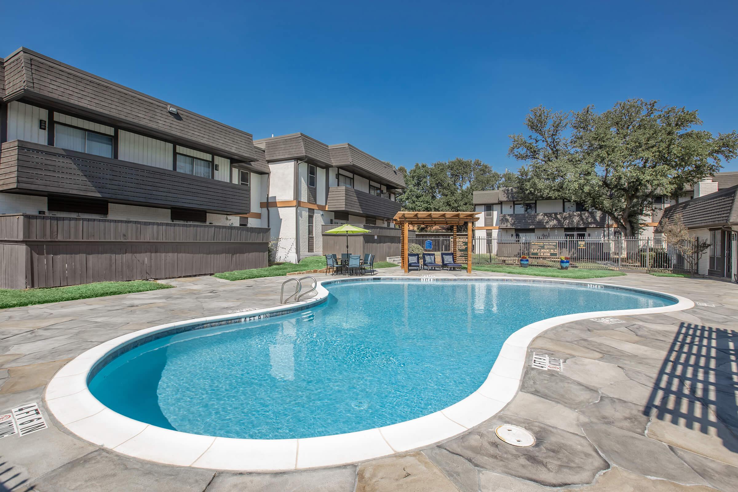 A clear swimming pool with a curved shape, surrounded by stone pavement and a wooden pergola with patio furniture. In the background, there are multi-story apartment buildings and well-maintained greenery under a bright blue sky. Perfect for relaxation and outdoor activities.