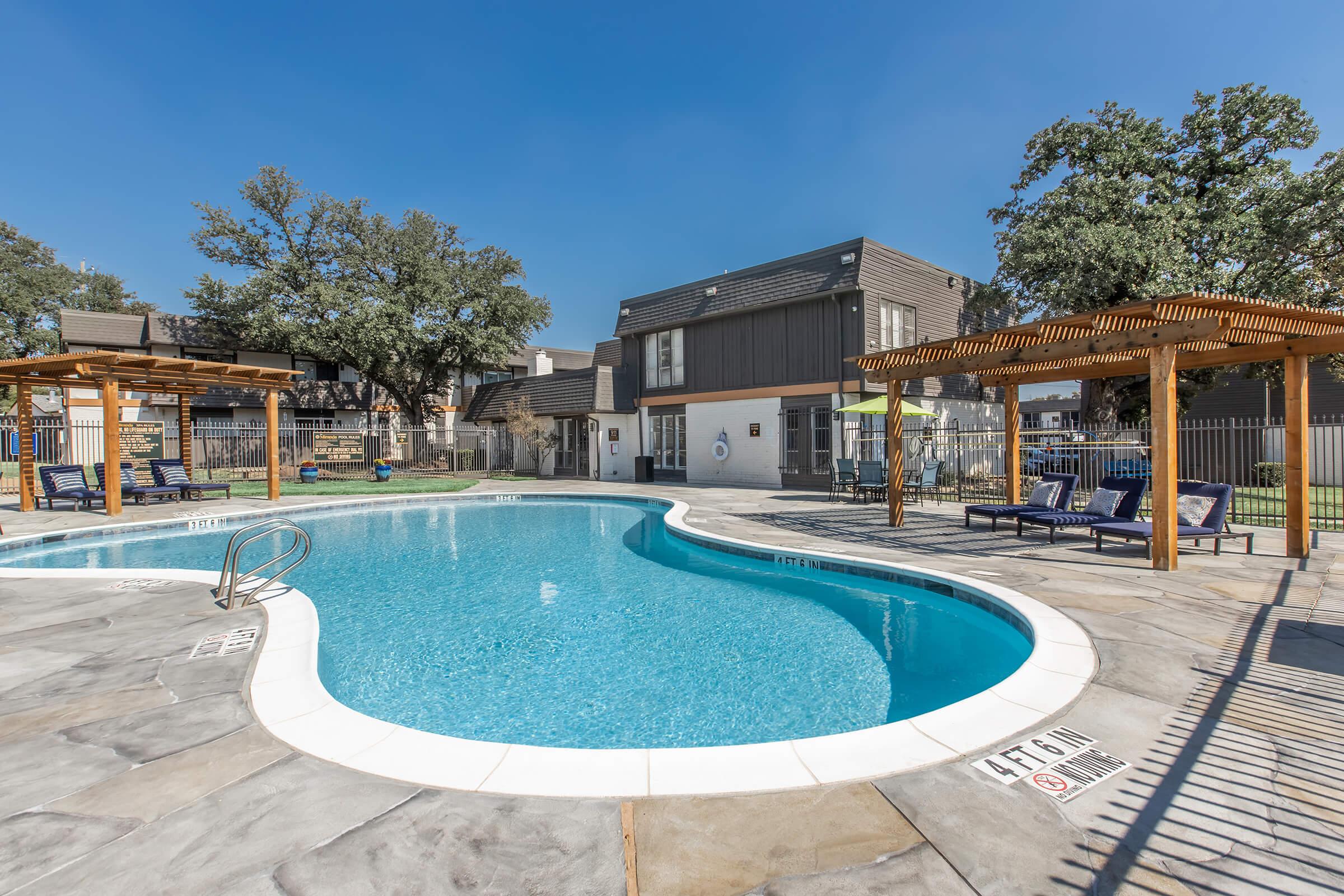 A clear blue swimming pool with a curved edge, surrounded by lounge chairs and wooden pergolas. In the background, a residential building with large windows and trees. The sky is bright and sunny, creating a cheerful atmosphere.