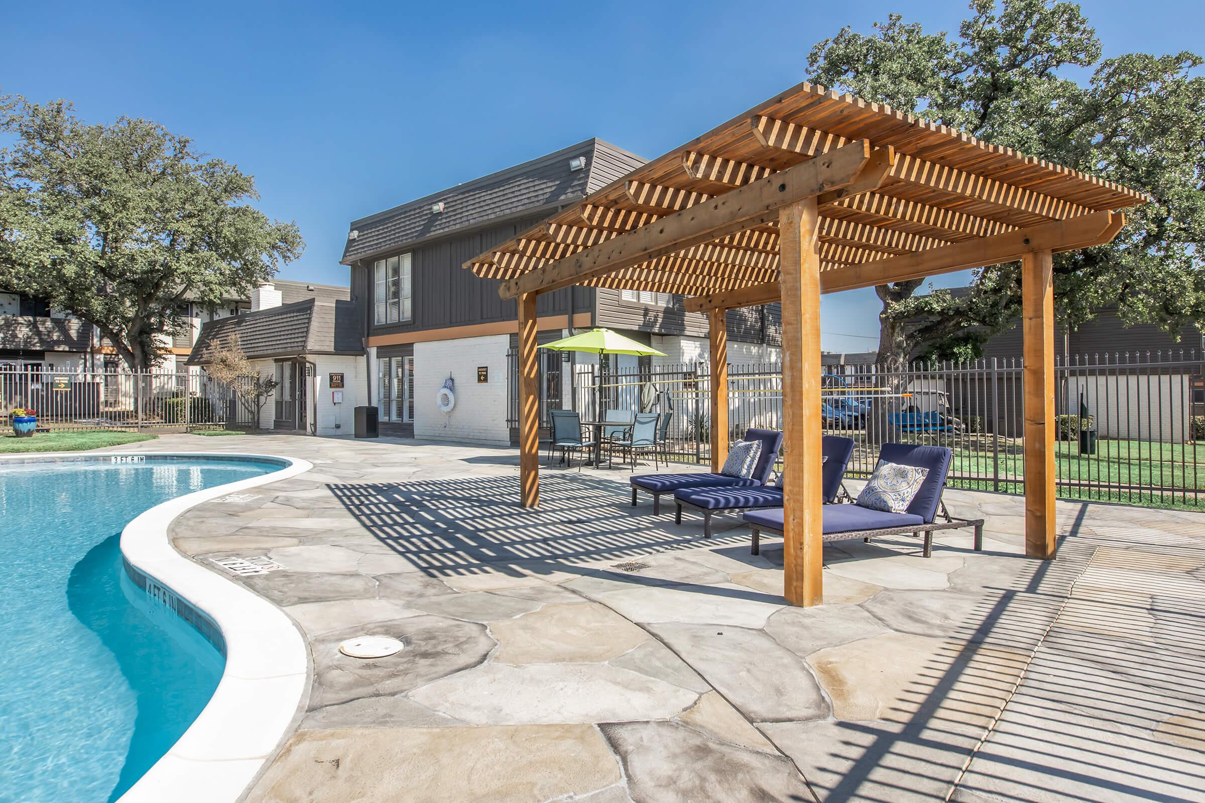 A swimming pool area featuring a wooden pergola, lounge chairs, and a sunny outdoor setting. The pool is surrounded by stone decking, with grass and buildings visible in the background. Shadows from the pergola create a patterned effect on the ground.