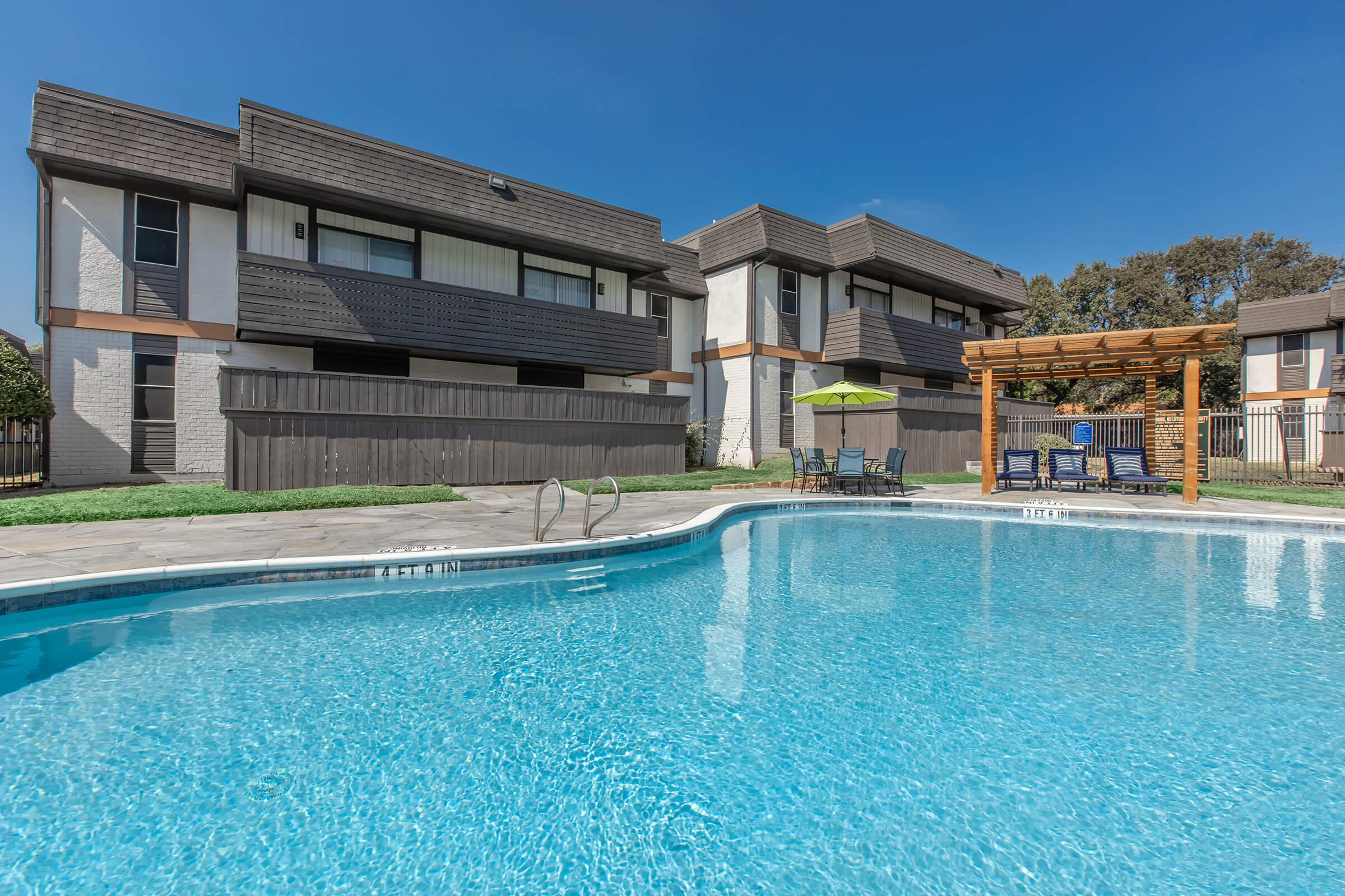 A clear blue swimming pool surrounded by a concrete deck, with lounge chairs and an umbrella nearby. In the background, two multi-story apartment buildings with brown roofs and a wooden pergola are visible, along with a well-maintained lawn. The sky is bright and blue.