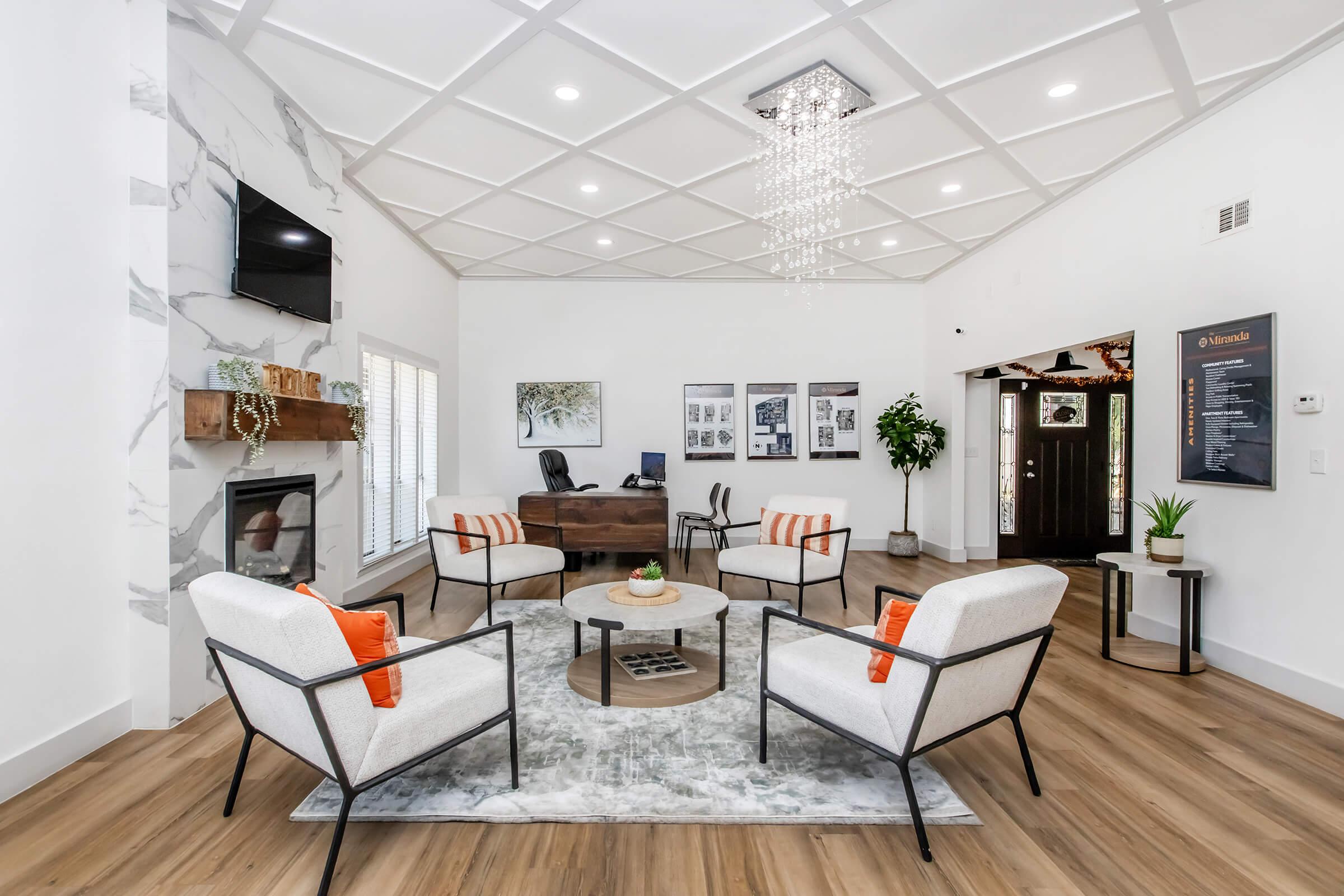 A modern living room featuring a bright atmosphere with white walls and coffered ceiling. The space includes a cozy seating area with four armchairs and an area rug, a round coffee table with a decorative centerpiece, a stylish fireplace, and a small desk in the background. Large windows add natural light.