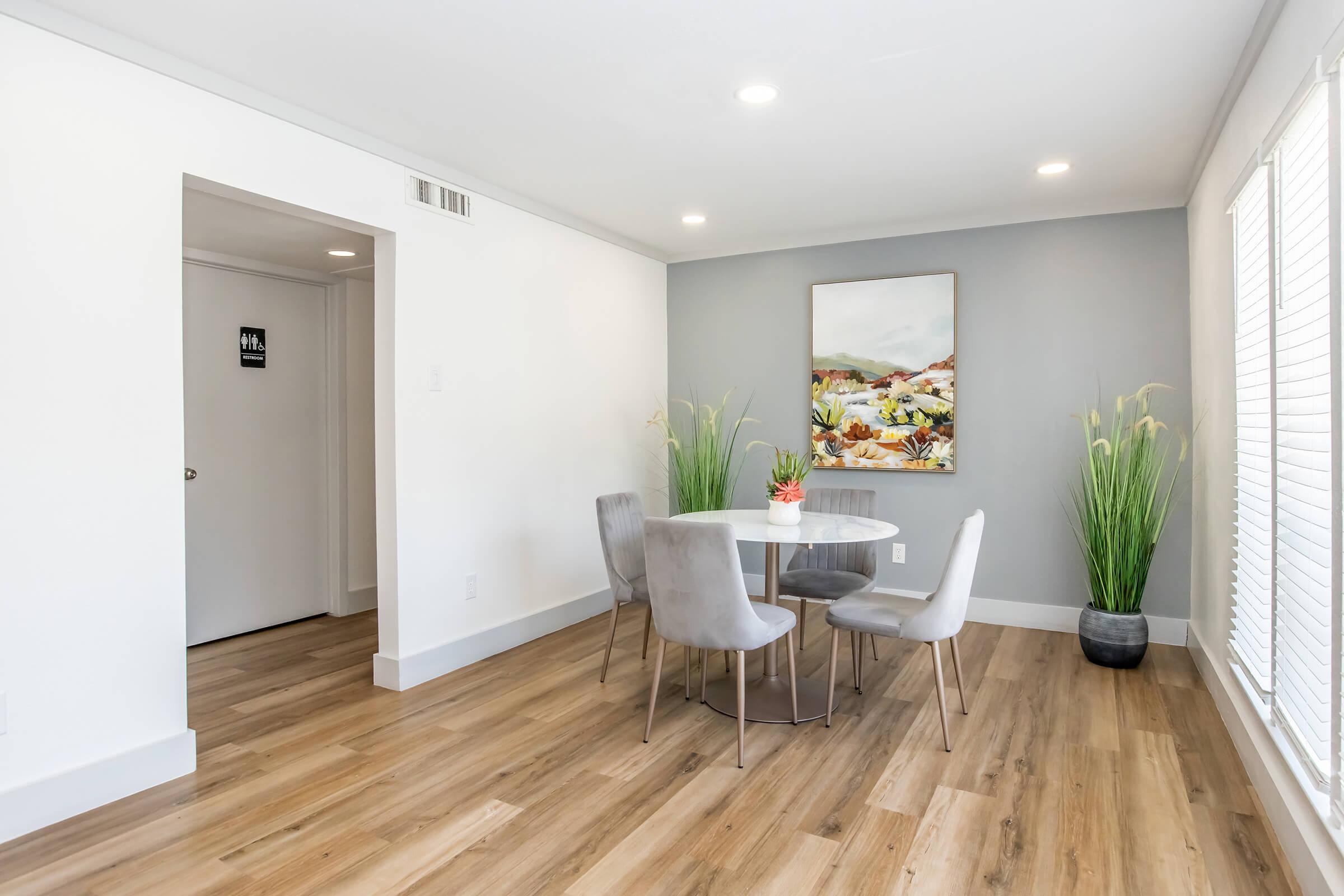A modern dining area featuring a round white table with four gray chairs. The space is decorated with a colorful abstract painting on the wall and two tall green plants in pots. There is a door on the left side of the image, and natural light streams in through the windows, enhancing the clean, contemporary design.
