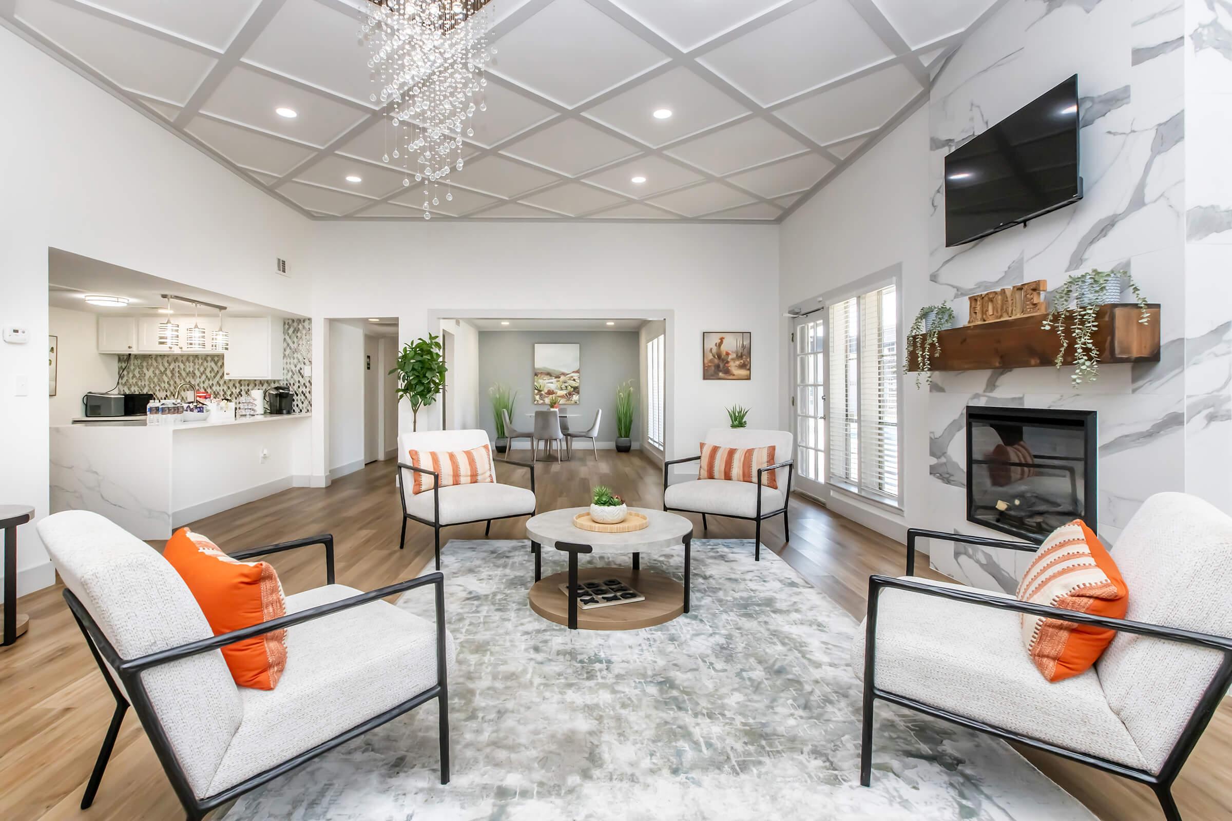 A modern, elegantly designed living room featuring a coffered ceiling, a stylish chandelier, and a cozy fireplace. Four armchairs with orange and white cushions are arranged around a circular coffee table on a decorative area rug. Large windows allow natural light, and a flat-screen TV is mounted on the wall.