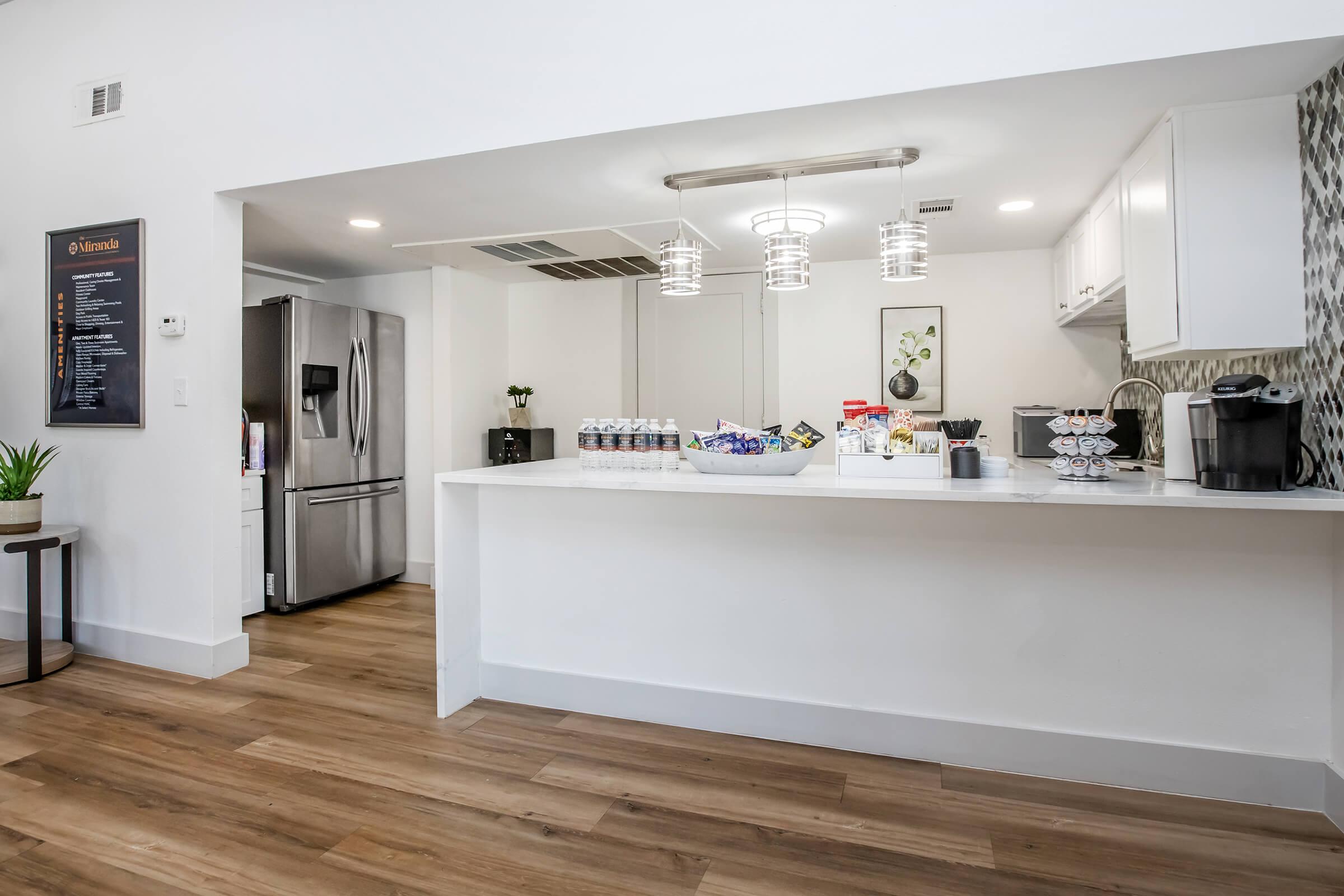 A modern kitchen space featuring a bar counter with snacks and drinks, including bottled beverages. A stainless steel refrigerator is visible, along with a coffee maker. Stylish pendant lighting hangs above the counter, and there are decorative elements like plants. The overall aesthetic is clean and inviting.