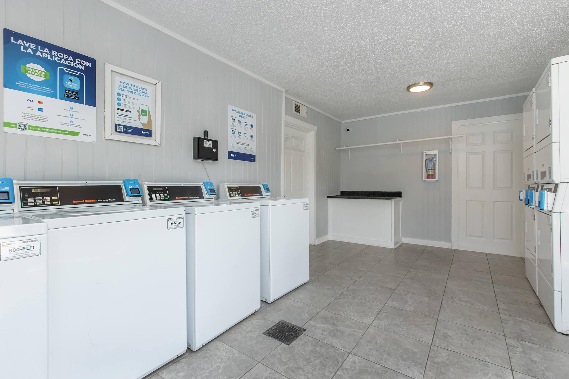 A laundry facility featuring several white washing machines and a countertop. The walls are light gray, and there are informational signs about laundry services. A door is visible at the back, with a notice board and a wall-mounted device near the entrance. The floor is tiled and has a drain.