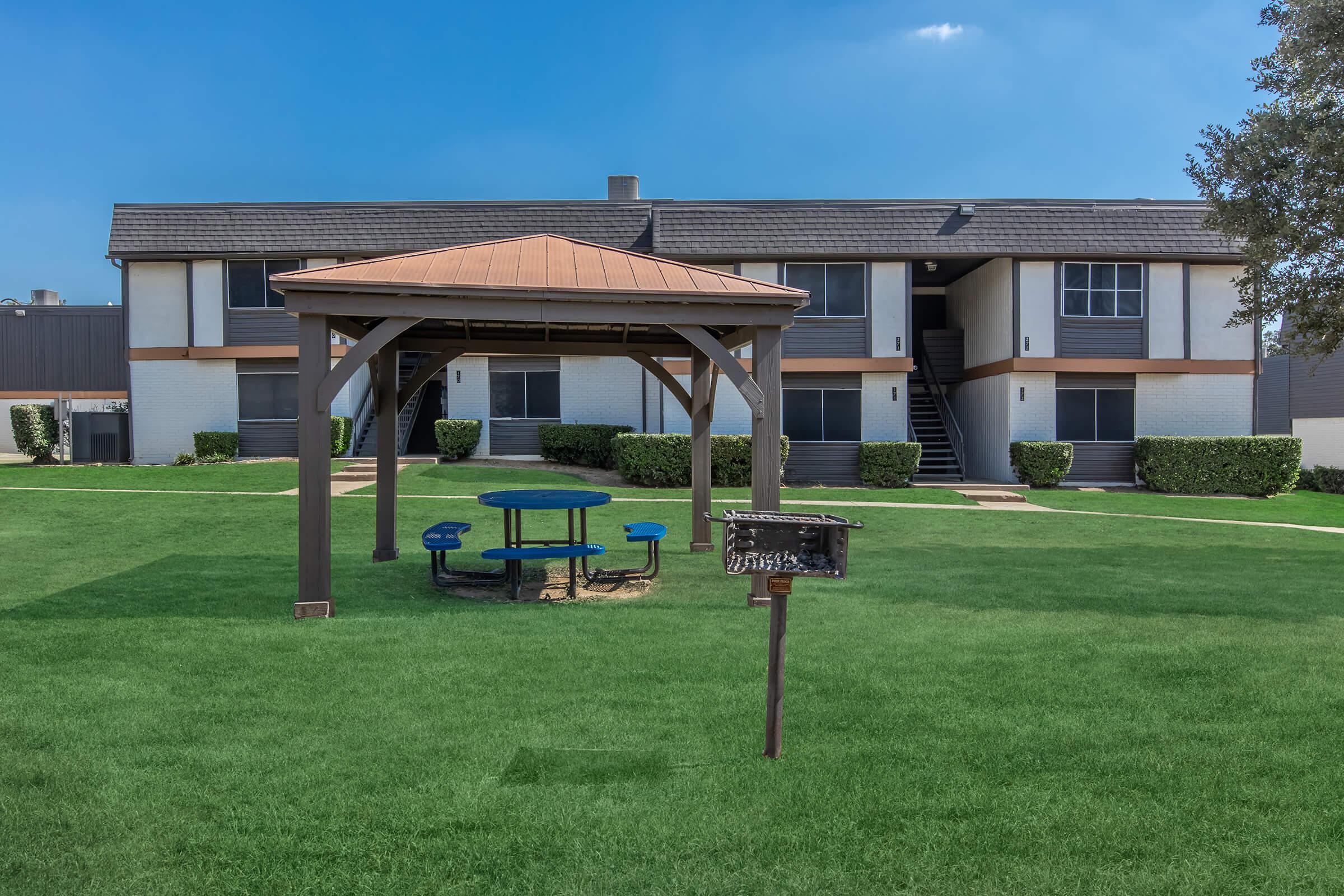 A well-maintained outdoor area featuring a gazebo with a brown roof, surrounded by green grass. Below the gazebo, there is a round blue picnic table with benches. In the background, a two-story apartment building with white and brown siding is visible, along with neatly trimmed shrubs and a clear blue sky.