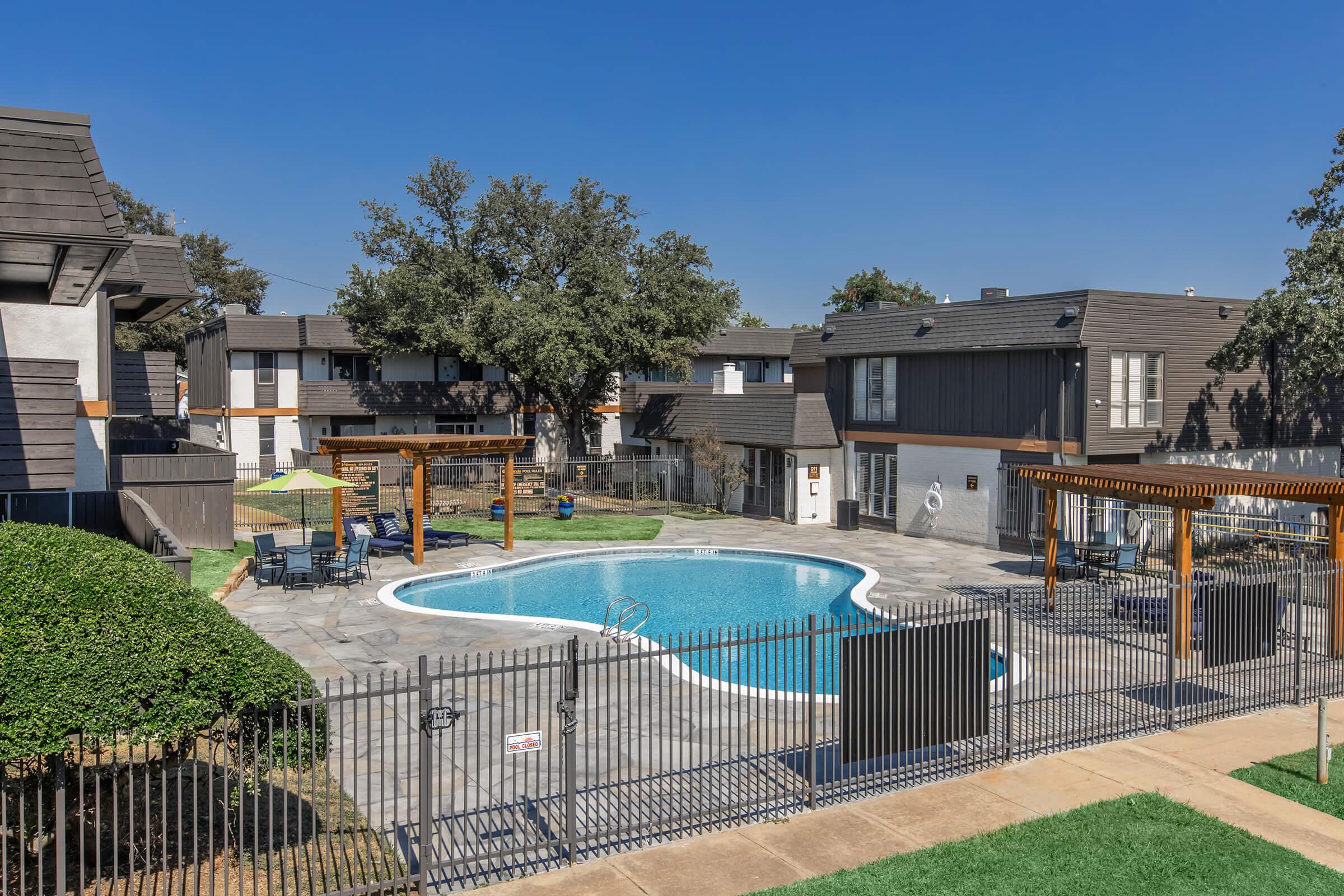 A well-maintained outdoor swimming pool surrounded by lounge chairs and shaded areas. The pool is enclosed by a decorative fence, and there are nearby apartment buildings with modern architecture and landscaping featuring grass and trees under a clear blue sky.