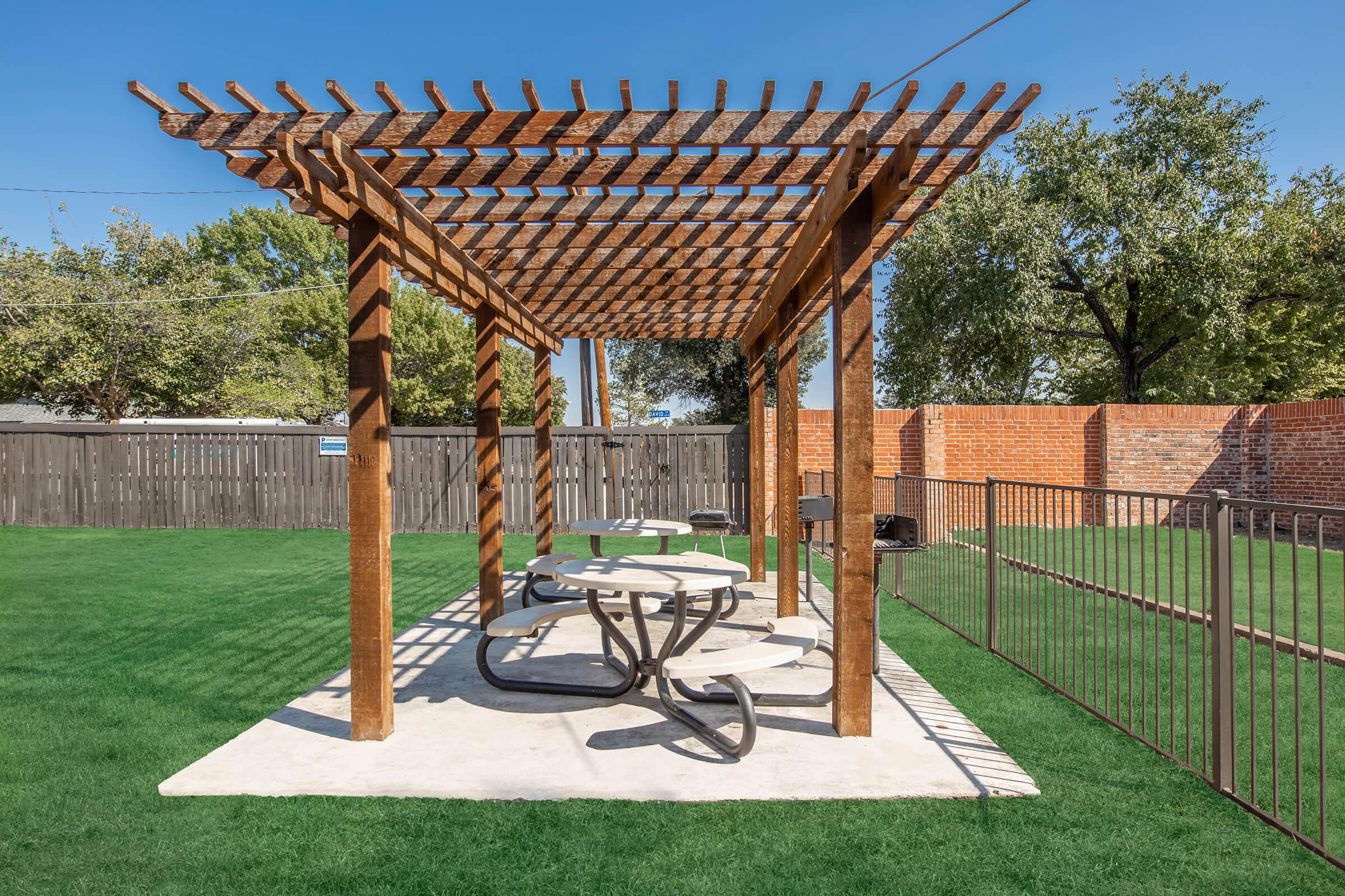 A wooden pergola with a slatted roof shades a concrete seating area featuring two circular tables with curved metal bases, situated on green grass. A wooden fence and brick wall are visible in the background, under a clear blue sky.