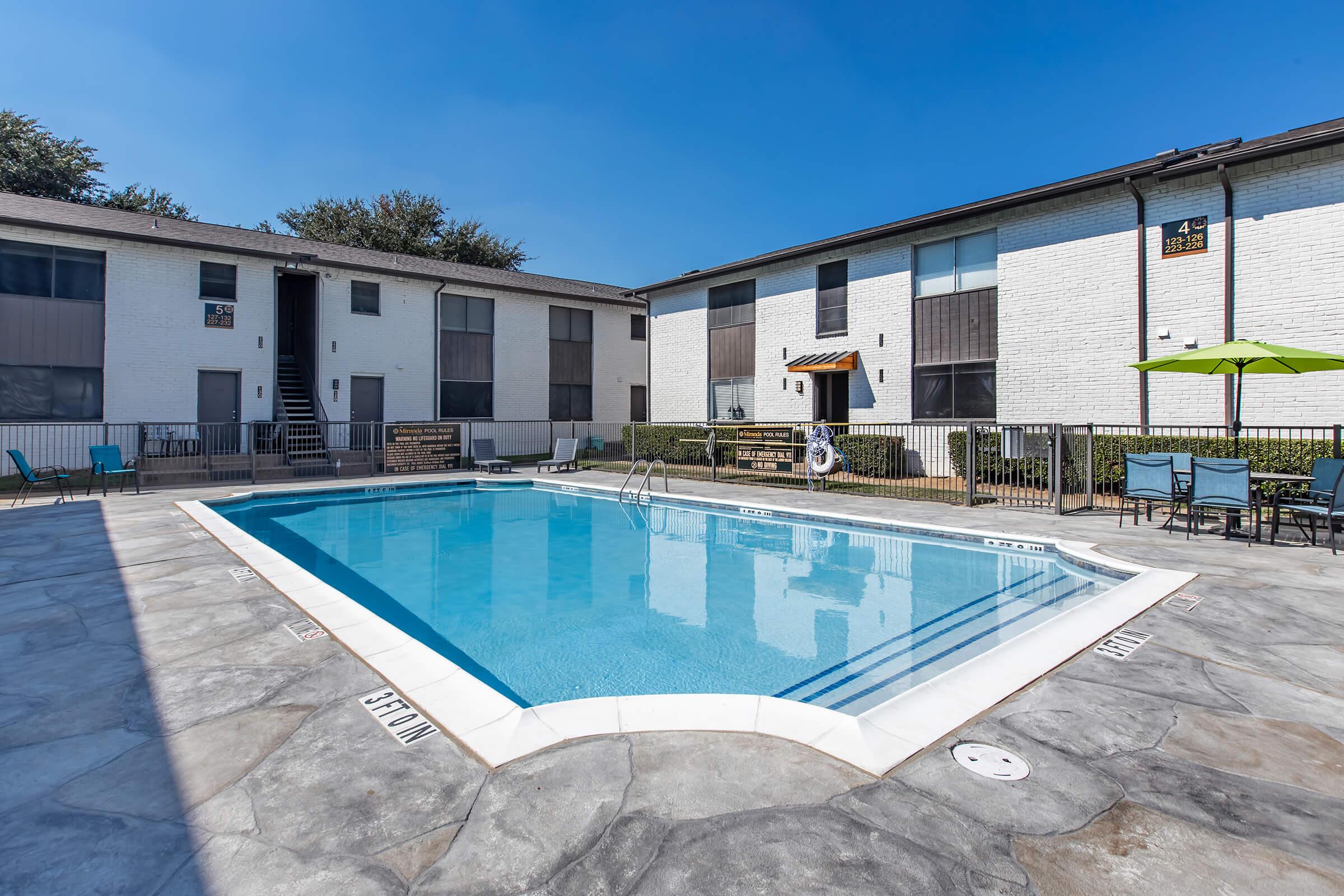 A clear blue swimming pool surrounded by a stone deck, with lounge chairs and green umbrellas nearby. In the background, two multi-story apartment buildings with white and dark exteriors, and a sunny sky with a few trees.
