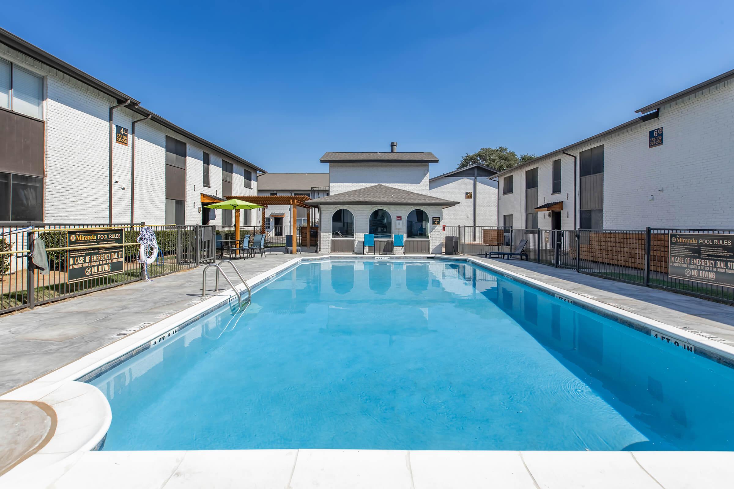 A clear blue swimming pool surrounded by a fenced area, with lounge chairs and an umbrella visible. In the background, there are residential buildings with white exteriors and green landscaping under a clear blue sky.