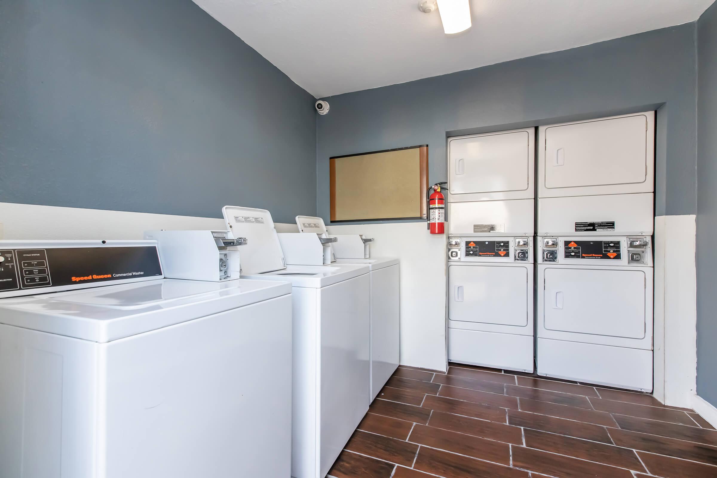 A clean, modern laundry room featuring several white washing machines and dryers. The space is well-lit with a gray wall and brown tiled flooring. A fire extinguisher is visible on the wall, and a mirror reflects part of the room. The overall ambiance is tidy and functional.