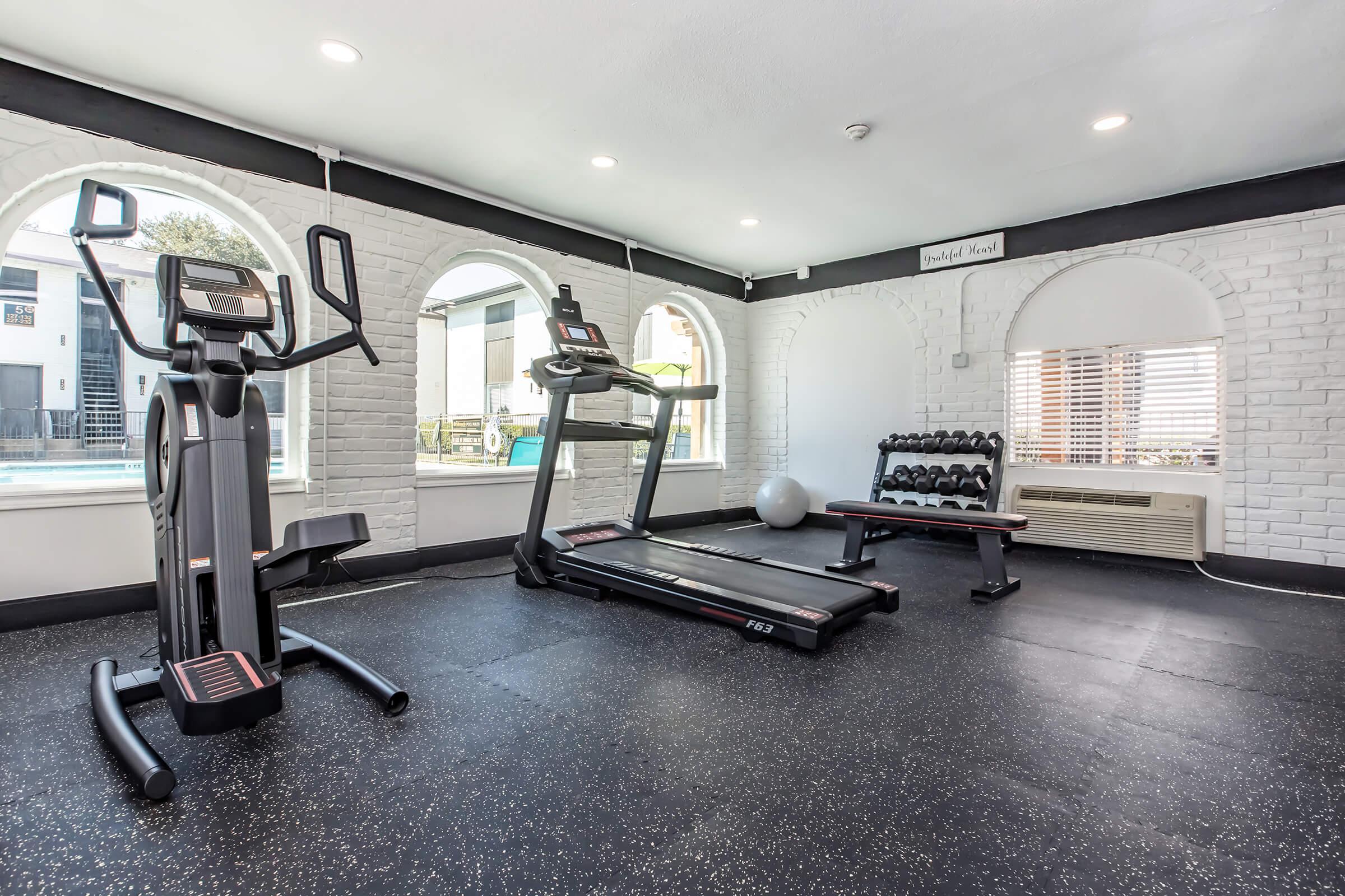 A modern gym featuring a treadmill, an elliptical machine, and a set of dumbbells on a rack. The room has large arched windows providing natural light, with a pool visible outside. The walls are bright white brick, and the flooring is black rubber.