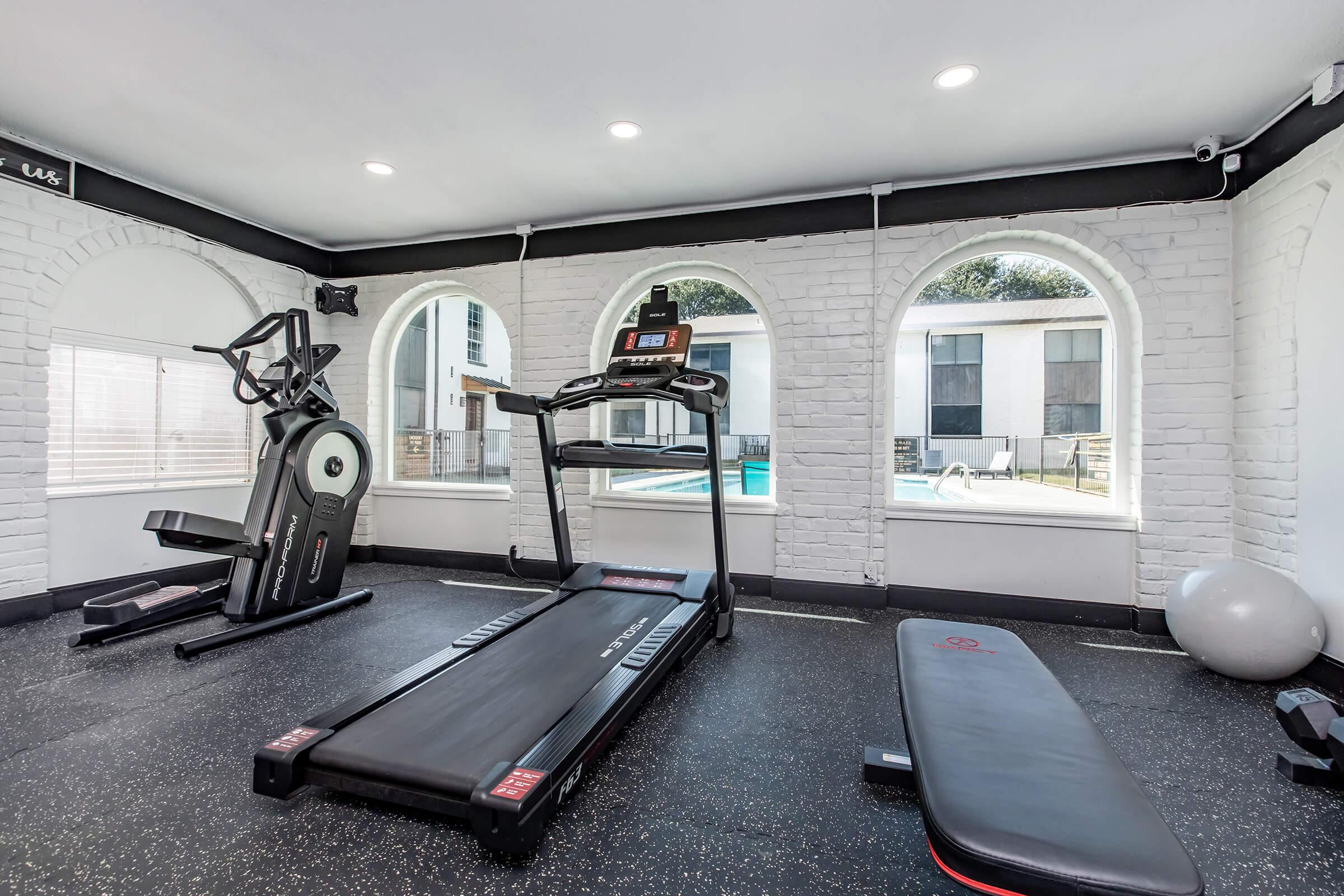 A modern gym interior featuring a treadmill, elliptical machine, and a flat bench on a rubber flooring surface. Large windows provide natural light and a view of an outdoor pool area. The walls are painted white with arched details, creating an inviting workout space.