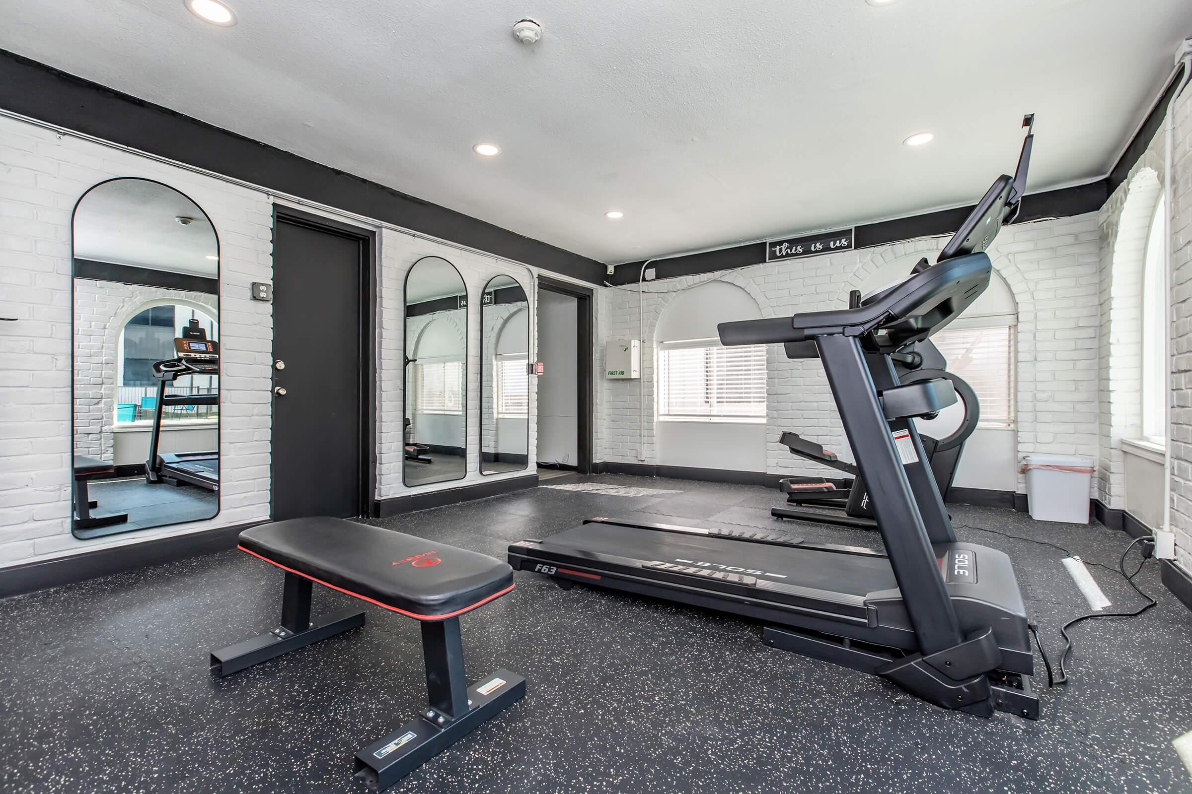A modern gym interior featuring a treadmill, a weight bench, and large mirrors against white brick walls. The space is well-lit, with large windows and a clean, minimalist design, creating an inviting atmosphere for workouts.