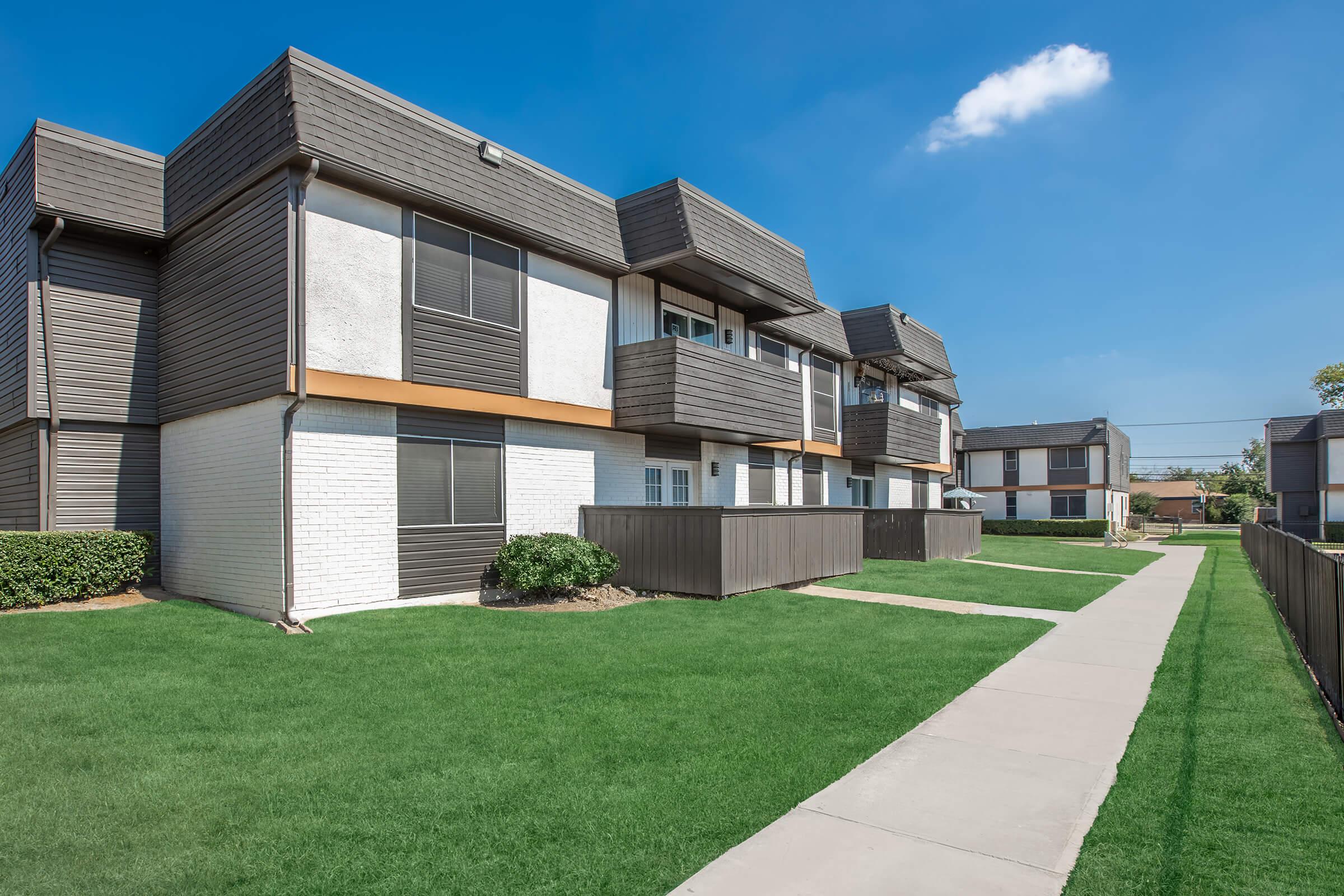 Multi-unit residential building featuring two-story design with balconies, a well-maintained lawn, and a clear blue sky. Sidewalks lead through the green grass, providing access to the units. The architecture showcases a modern style with a mix of brick and siding materials.