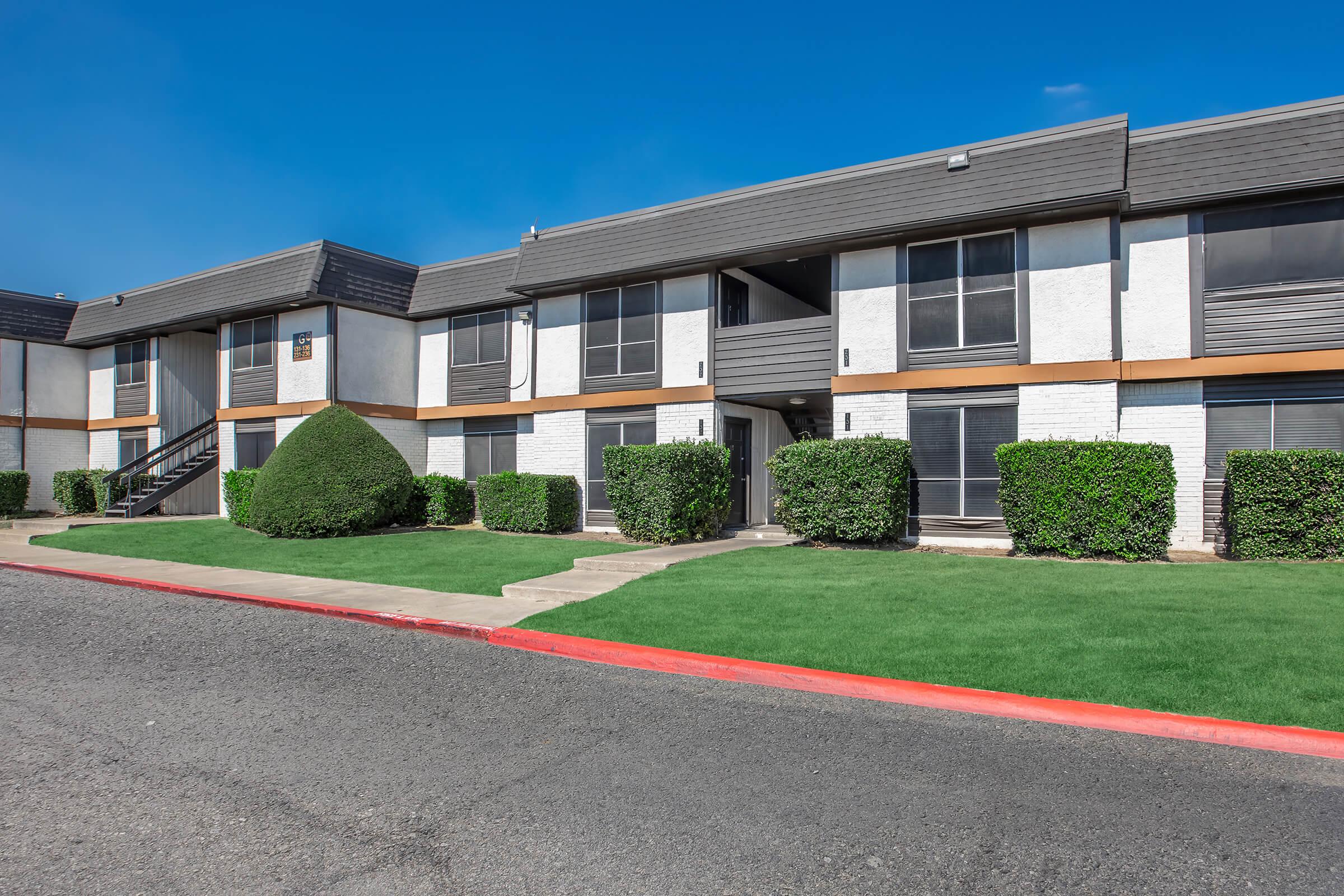 A modern apartment complex featuring two-story buildings with a neatly landscaped front yard, including manicured bushes and green grass. There are dark gray roofs with large windows, and a concrete pathway leading up to the entrances. The sky is clear and blue, contributing to a bright and inviting atmosphere.