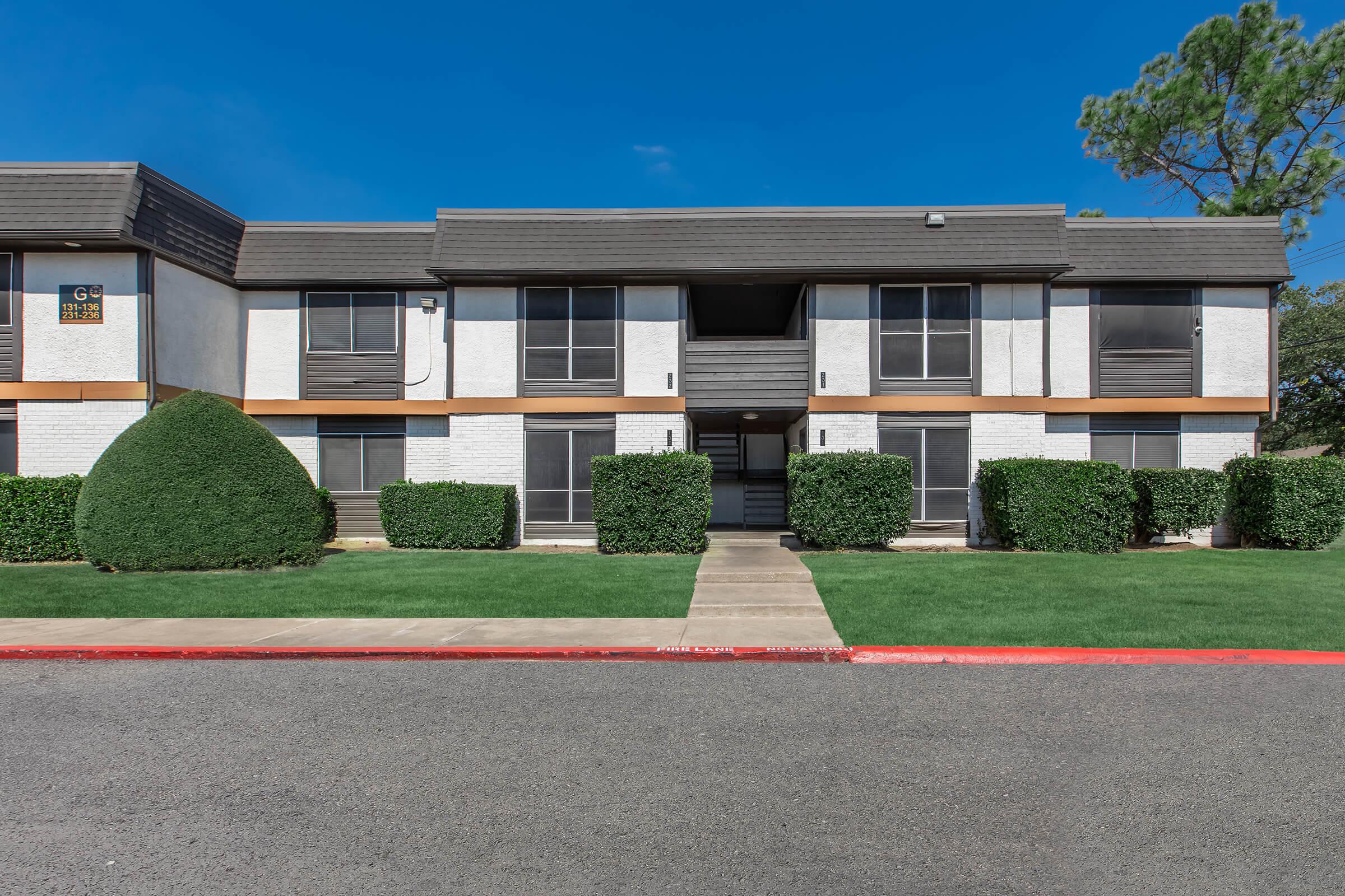 A modern two-story apartment building with a light-colored facade and dark trim, flanked by neatly trimmed hedges and a green lawn. The entrance features a set of stairs leading to the upper floor. The sky is clear and blue, adding to the bright appearance of the scene.