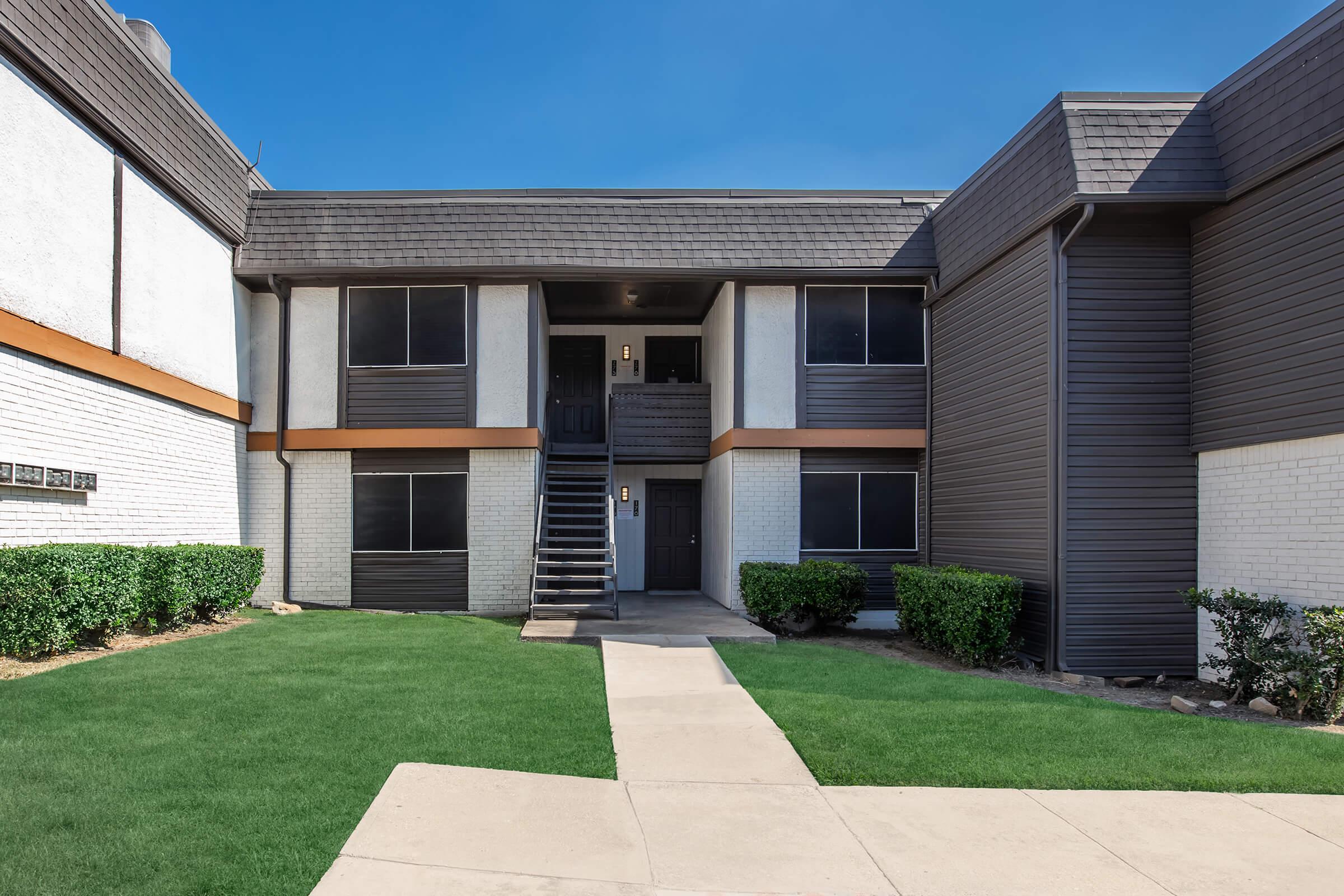 Two-story apartment building with a modern design featuring a dark exterior, multiple windows, and a staircase leading to the upper units. The surrounding area has well-manicured grass and light-colored pathways, creating a welcoming atmosphere. Clear blue sky in the background.
