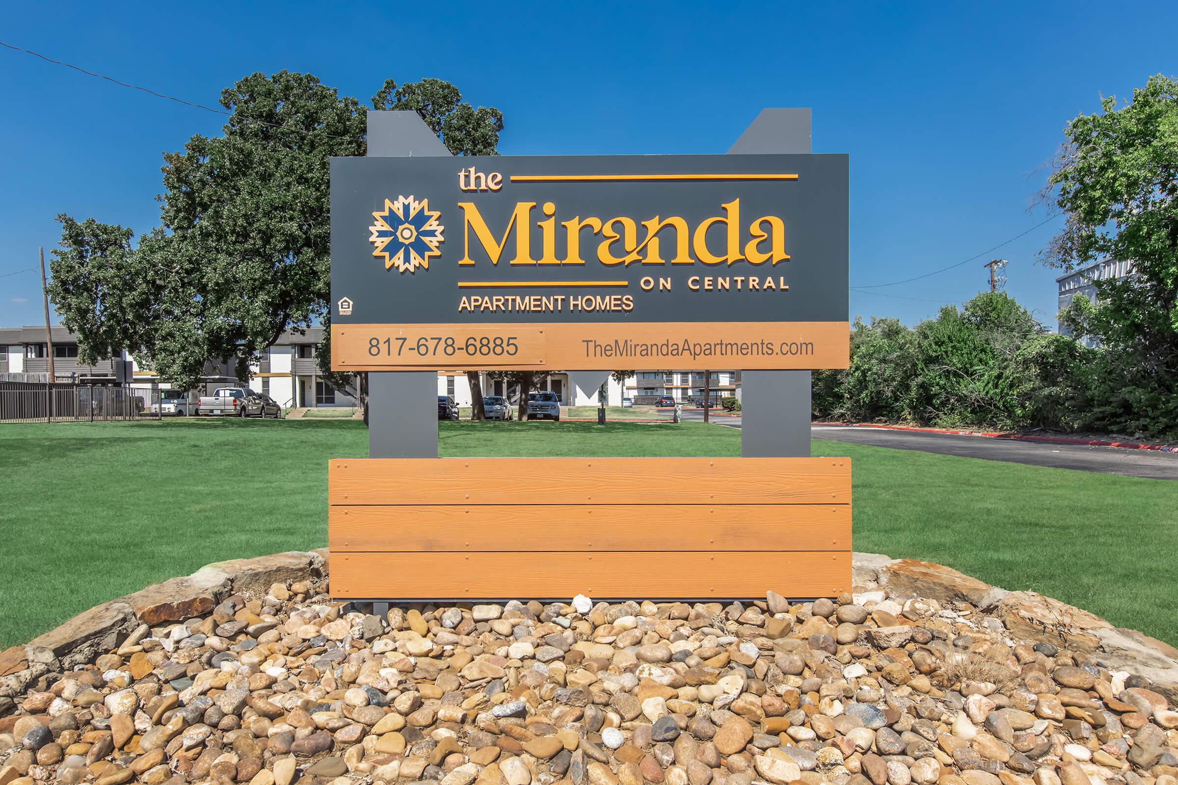 Sign for "The Miranda on Central Apartment Homes" with contact number 817-678-6885. The sign features a sun logo and is placed on a landscaped area with rocks in the foreground and greenery in the background.