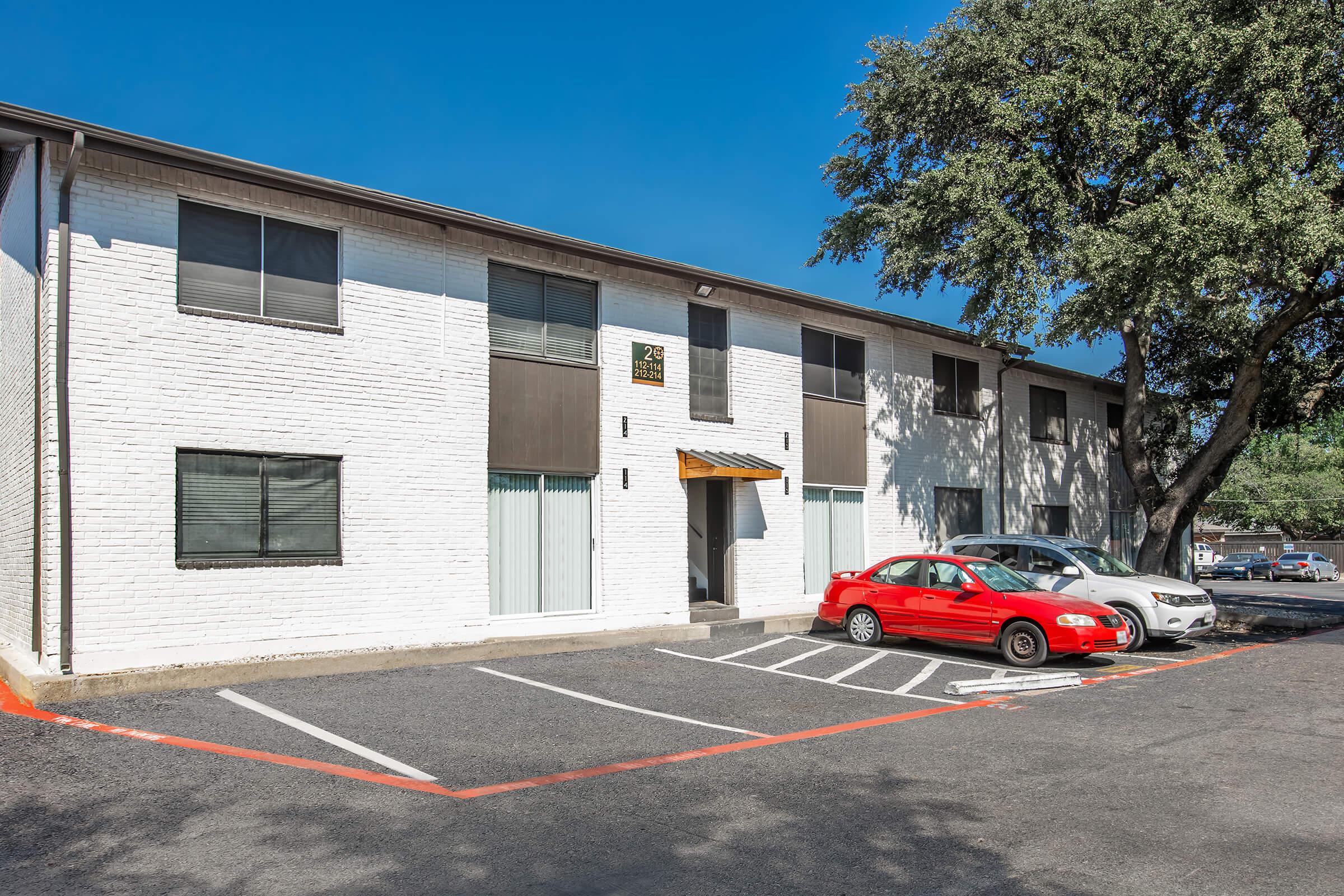 A two-story residential building with a white brick exterior, featuring large windows and a wooden canopy over the entrance. A red car is parked in front, along with another vehicle. The scene is set under a clear blue sky with a tree providing shade in the foreground.