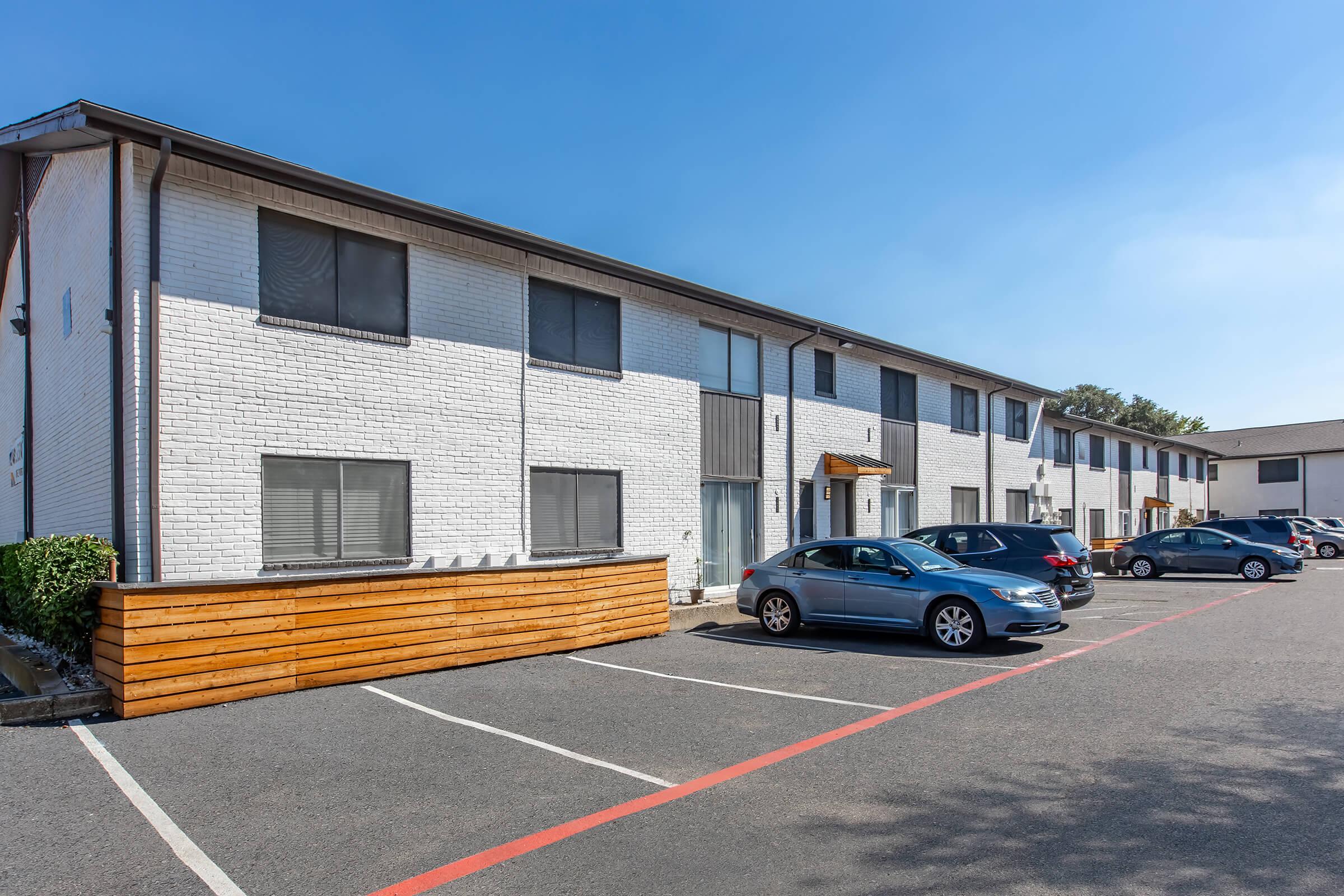 A view of a multi-story apartment building with a white brick exterior. There are several parked cars in front, and a wooden barrier on one side. The sky is clear and blue, providing a bright atmosphere. The building features multiple windows and a partially shaded entrance.