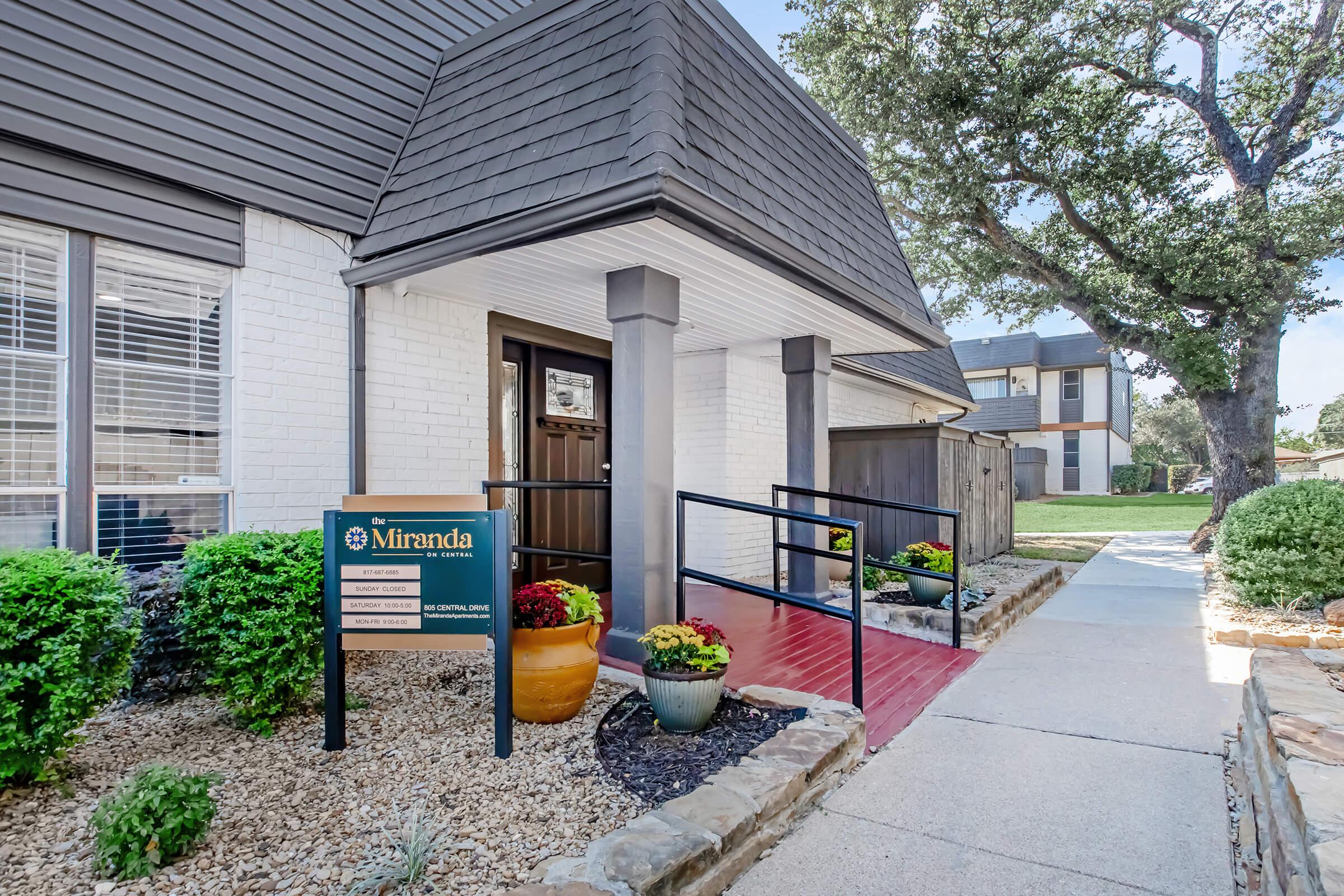Exterior view of a building named "Miranda" with a welcoming entrance. The pathway features decorative potted plants and a ramp for accessibility. The building has a modern design with large windows and is surrounded by manicured landscaping. Trees are visible in the background, contributing to a pleasant environment.