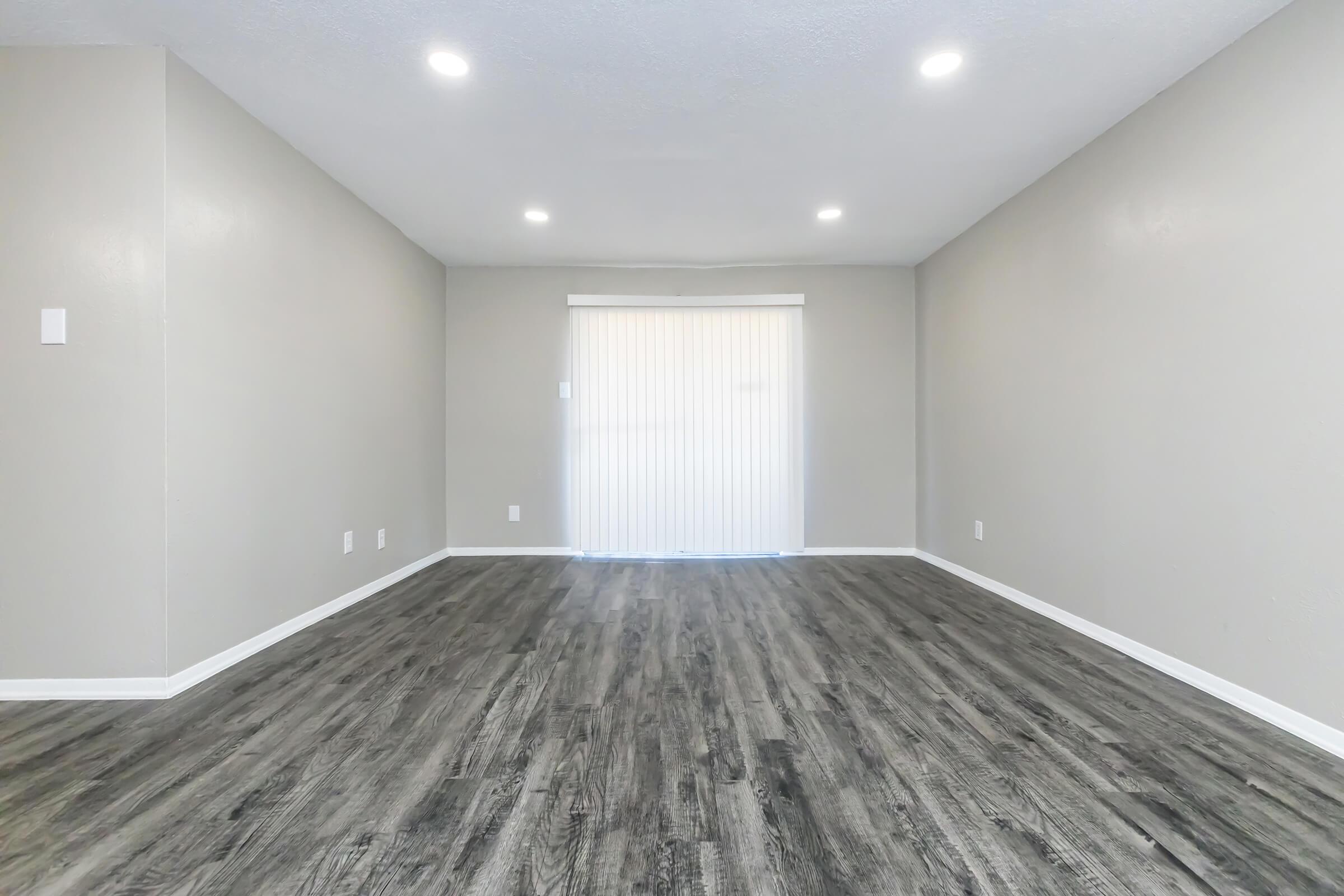 Empty interior room with light gray walls and laminate flooring. Brightly lit by recessed lights and a sliding door with vertical blinds at the far end, leading outside. The space has a minimalist feel, with no furniture or decorations present.