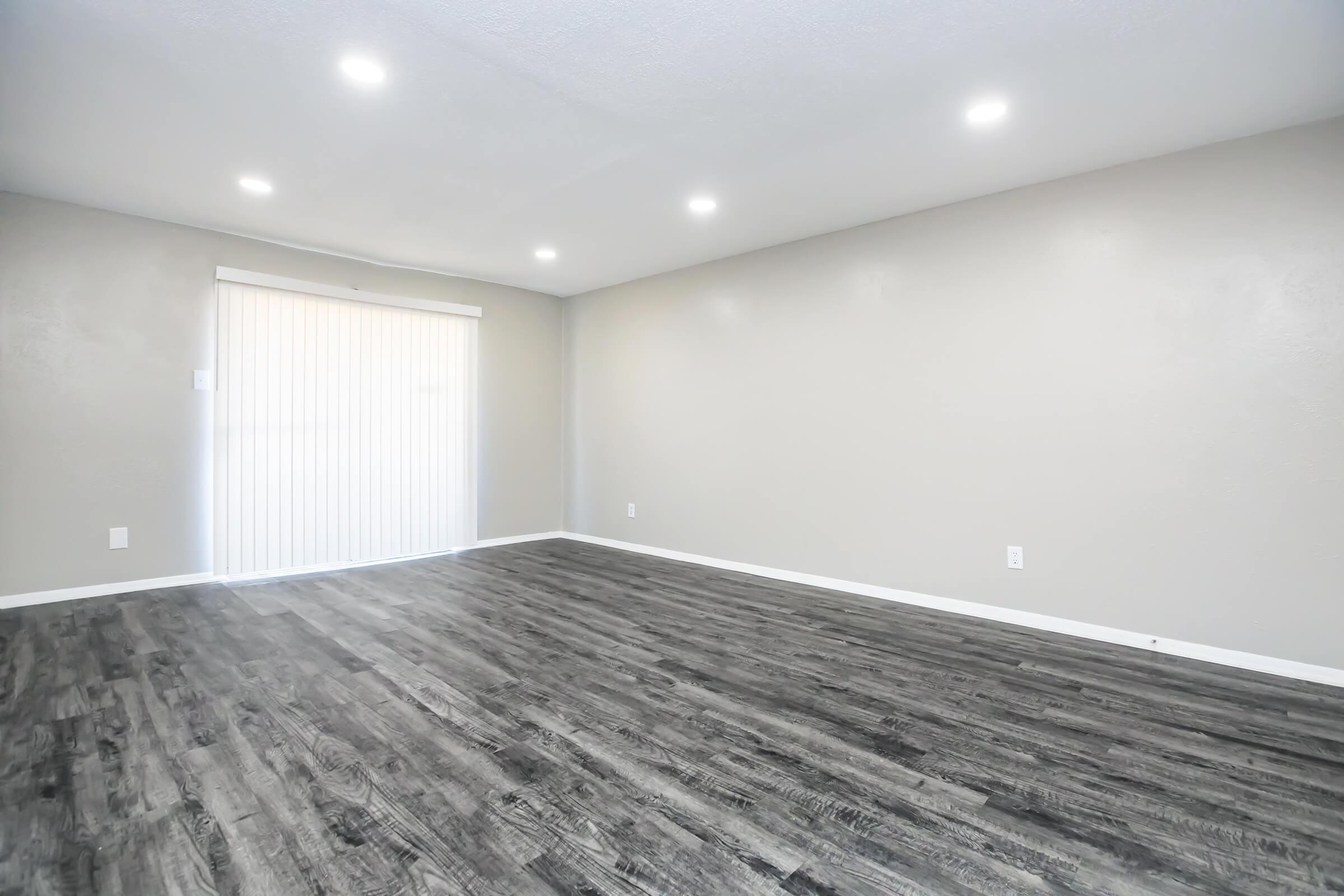 Empty room with gray walls and a large window covered by vertical blinds, featuring a modern laminate floor in dark wood tone. The ceiling is equipped with recessed lighting, creating a bright and spacious appearance.