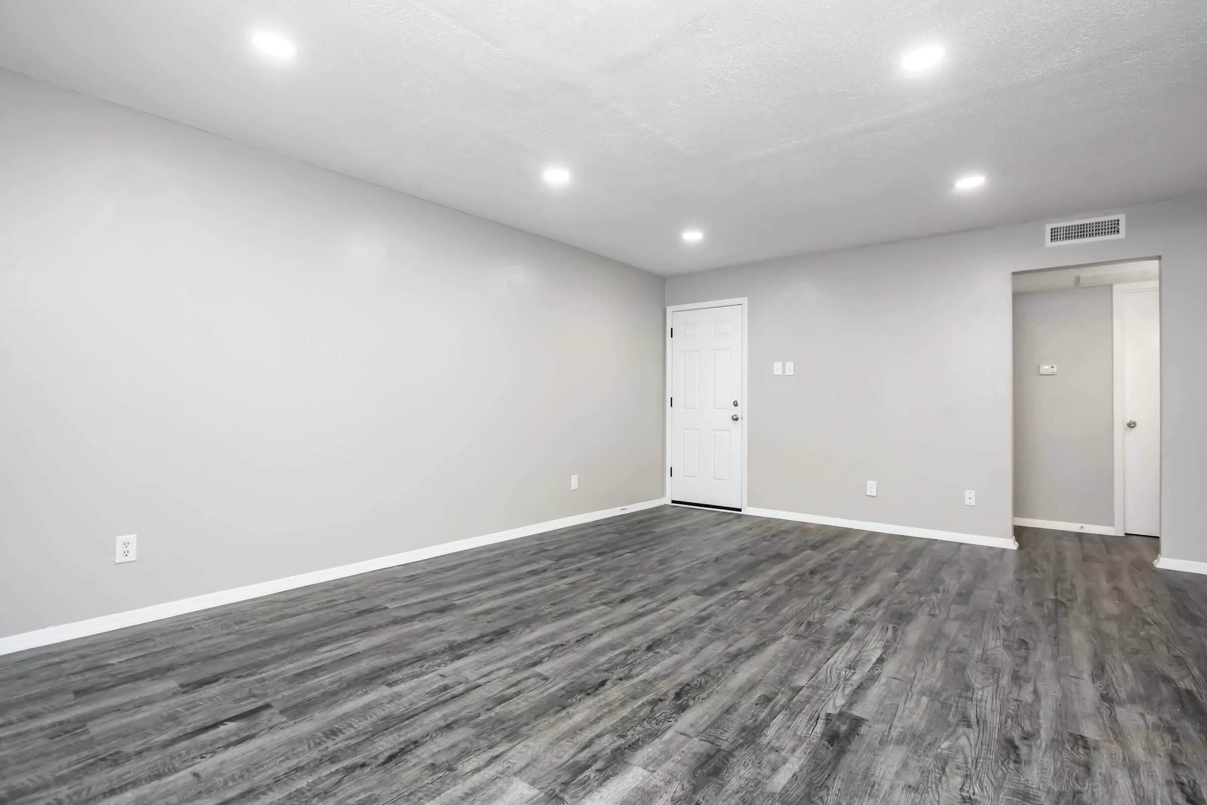 A spacious, empty room with light gray walls and dark wood-like flooring. The ceiling is equipped with recessed lighting, providing a bright ambiance. A white door is visible on the right side, leading to another room or hallway. The space appears clean and ready for furnishing.