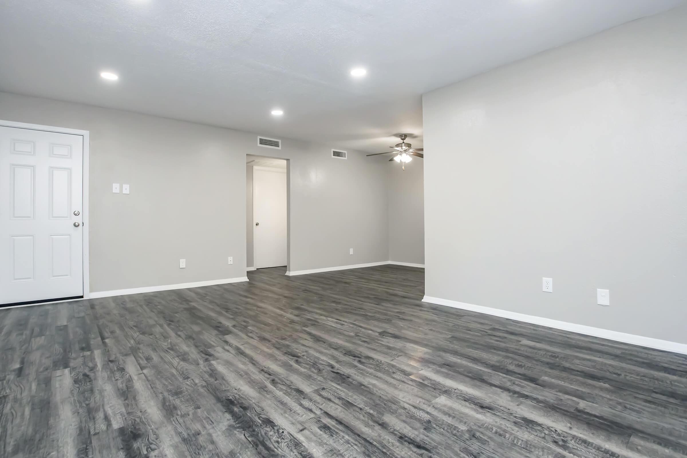 A spacious, modern living room with gray walls and dark wood-style flooring. The area features a ceiling fan, bright recessed lighting, and a front door on the left. A doorway leads to another room in the background, creating an open and inviting atmosphere.