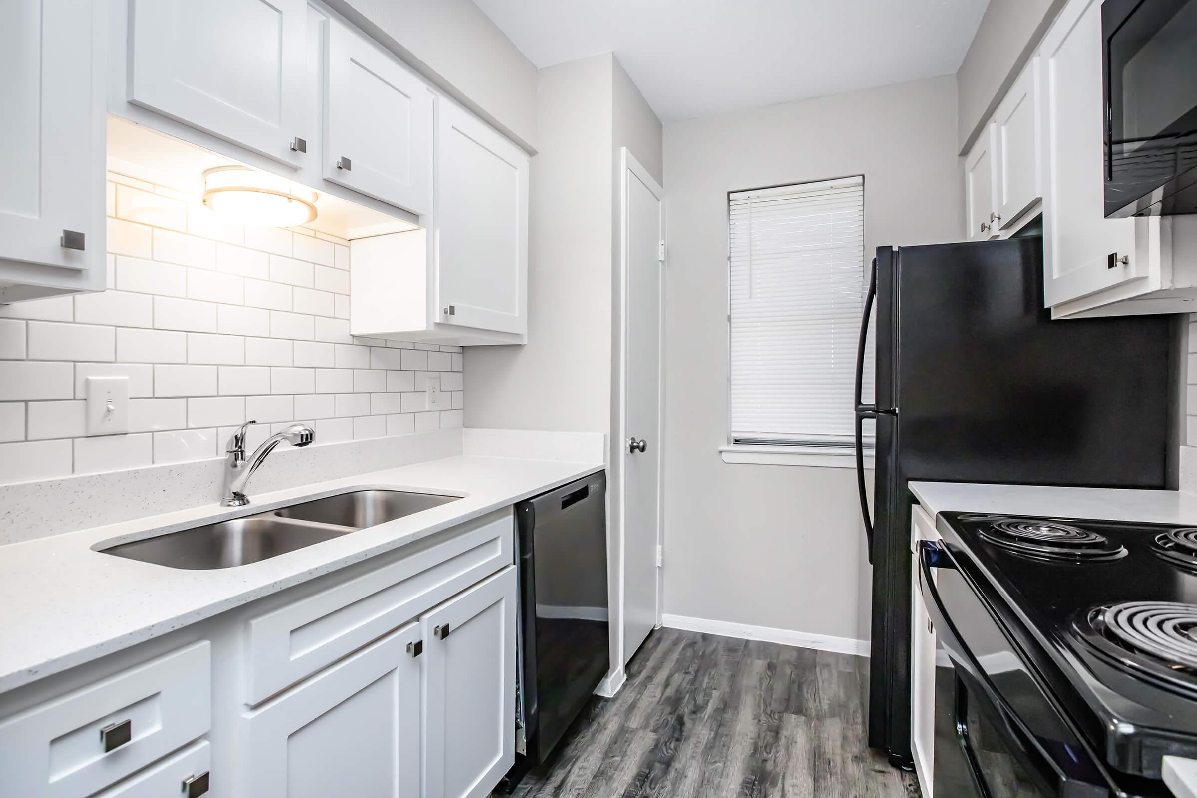 A modern kitchen with white cabinetry, laminate countertops, and stainless steel appliances. The features include a double sink, a dishwasher, a black refrigerator, and a black stove with an oven. The walls are painted light gray, and there is a small window with blinds.